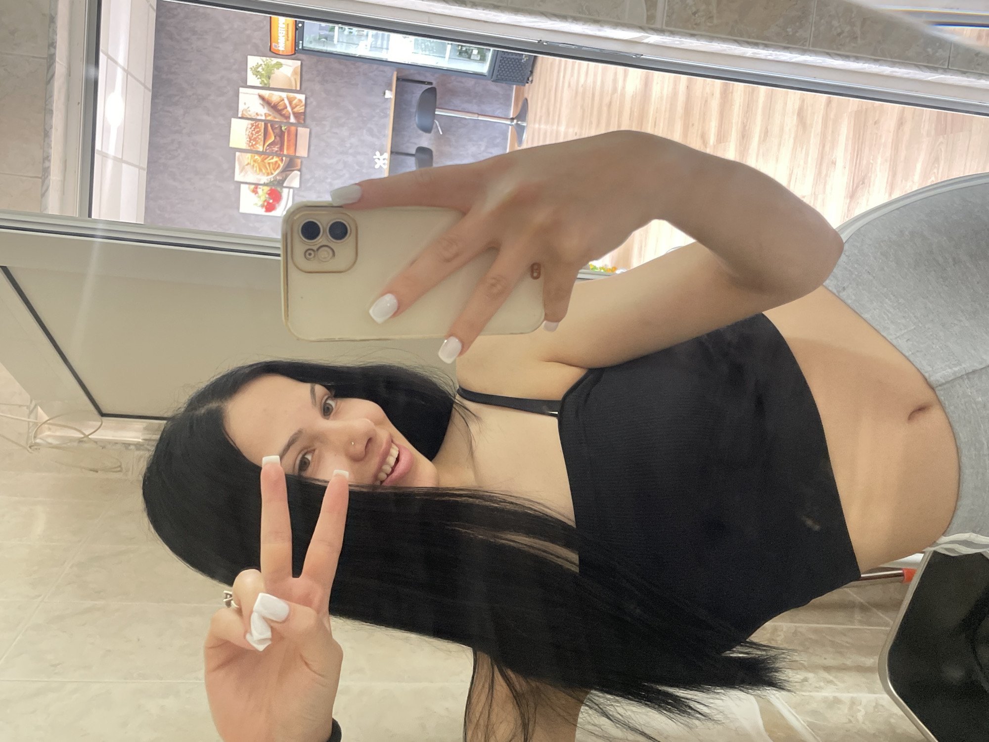 A young woman with long, dark hair is taking a mirror selfie. She is wearing a black crop top and grey sweatpants. Her nails are painted white. She is making a peace sign with her right hand and holding a smartphone in her left hand. In the reflection of the mirror, a food establishment with a counter and stools is visible. This scene is located in Stara Zagora, Bulgaria.