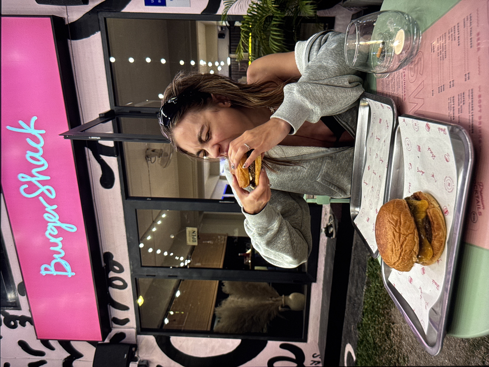 An outdoor shot shows a young woman eating a burger in front of a pink neon sign that reads "Burger Shack." The woman is wearing a grey hoodie and has a black headband on. She is sitting at a green table with two metal trays, one with a burger on it and the other empty. There is a glass candle holder with a lit candle and a cigarette butt in it on the table. In the background, there are glass doors with lights visible inside. The building is decorated with black graffiti. The scene takes place at night, suggesting an urban outdoor dining setting.