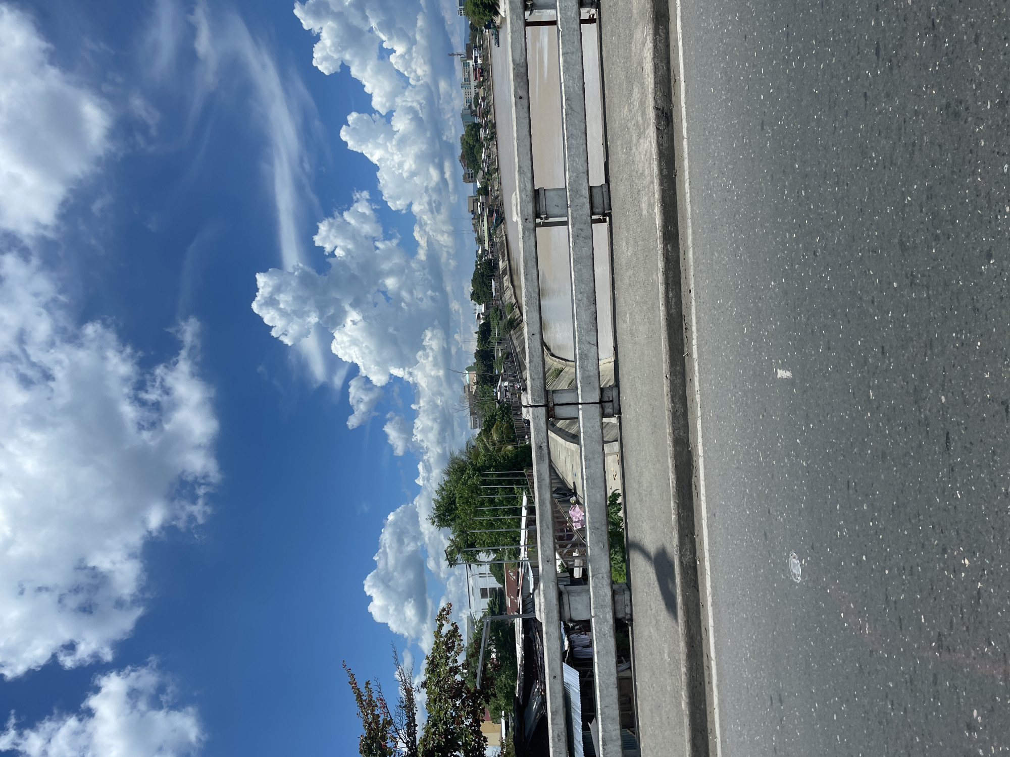 This is a photograph taken from a bridge in Davao City, Philippines, overlooking a river and the cityscape. The sky is bright blue with large, fluffy white clouds. On the left side of the frame, there are trees and some modest structures, including what appear to be corrugated metal roofs. The river is wide and appears to be muddy brown in color. Along the far bank of the river, a dense collection of buildings and some greenery are visible, suggesting a populated urban or semi-urban area. The bridge railing, made of concrete and metal bars, is a prominent feature in the foreground. The road surface is asphalt, with a visible curb and white road markings. No people are clearly discernible in the image, and no specific actions or interactions are depicted. The overall impression is a sunny day in a riverside city.