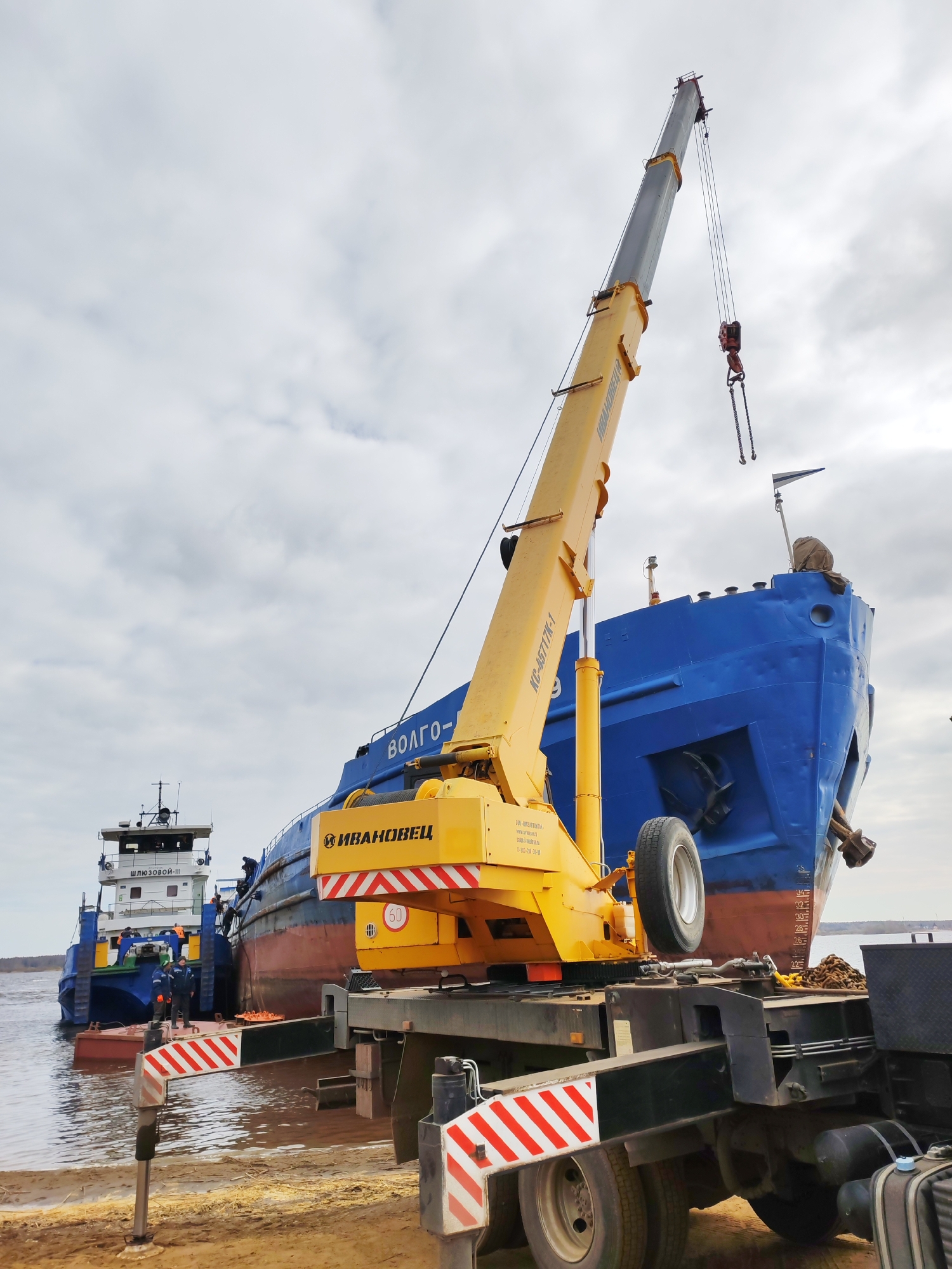 A yellow and black crane, operated by the company "Ivanovets," extends its boom towards a large, blue vessel named "Volgo-" on the shores of a body of water in Rybinsk, Russia. A smaller, white and blue ship with the name "Shlyuzovoy-III" in Cyrillic is docked beside the larger ship. The crane sits on a truck bed and has its stabilizers extended with red and white striped markings. The background is dominated by an overcast sky.