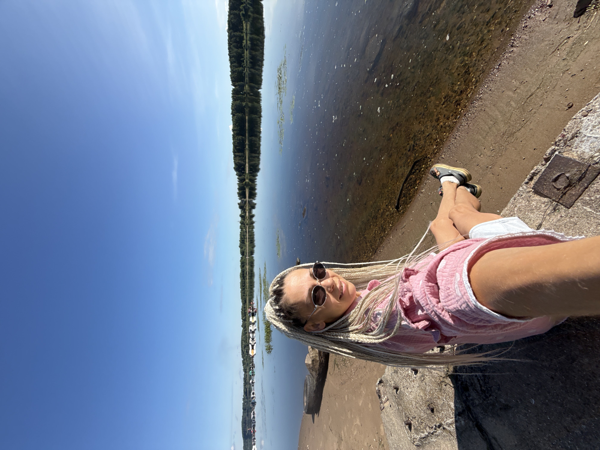 In Rusanovo, Russia, on a clear day, a woman with blonde braided hair and sunglasses sits on a concrete embankment next to a calm body of water. She wears a pink textured shirt and white shorts. The water reflects a line of trees on the opposite bank and a clear blue sky. Several boats are moored in the distance. The foreground shows the sandy and rocky shoreline.
