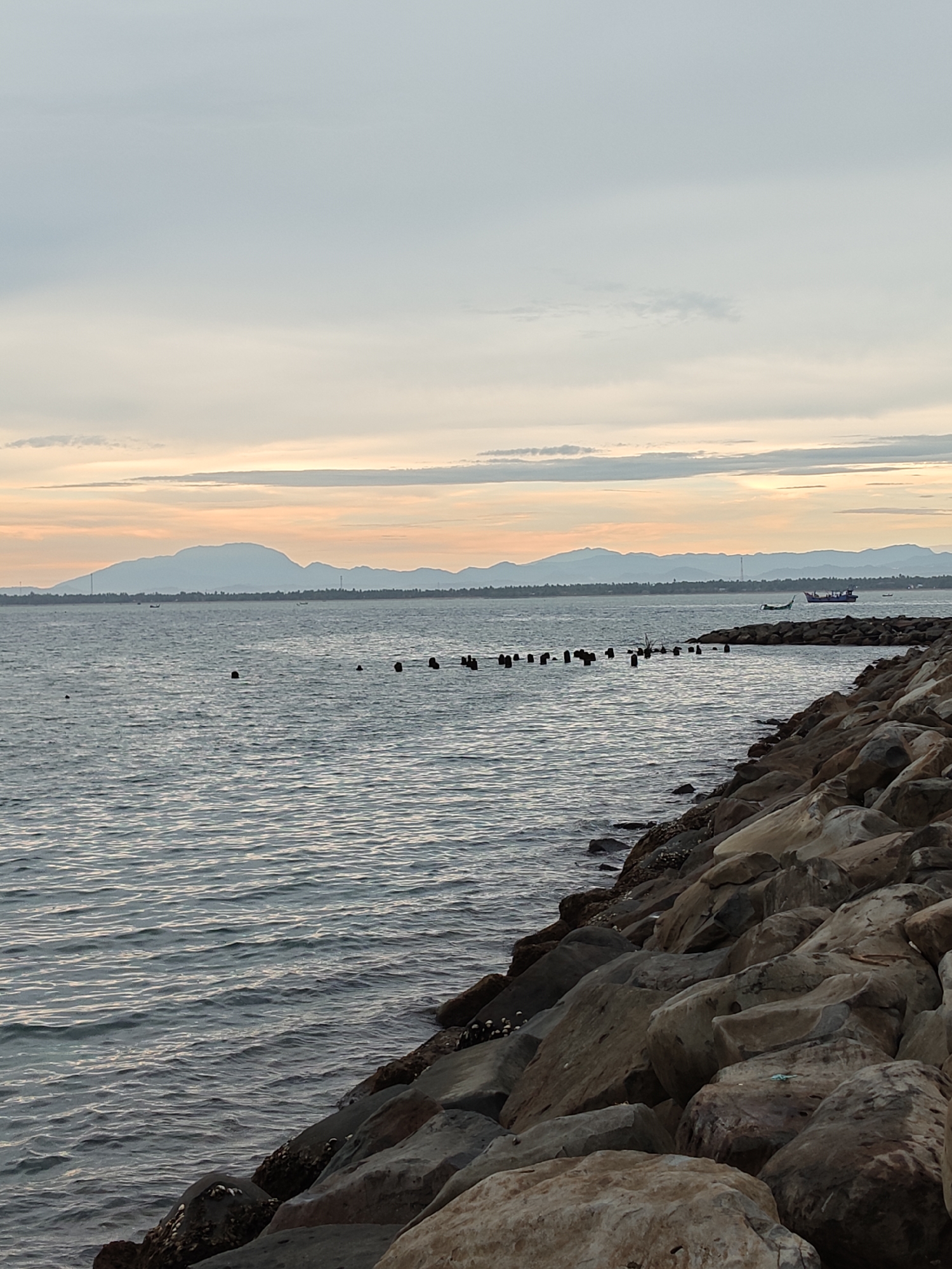 The image depicts a seascape in Lhokseumawe, Indonesia, at what appears to be either dawn or dusk, given the soft pastel hues in the sky. The horizon line features a range of distant mountains. The sea itself is calm, with gentle ripples on the surface.
In the midground, there's a row of weathered, vertical wooden posts extending into the sea, likely remnants of an old structure. A boat is visible on the water.
In the foreground, a rock barrier lines the coast. The rocks are large, varying in size and color, with some covered in marine growth.