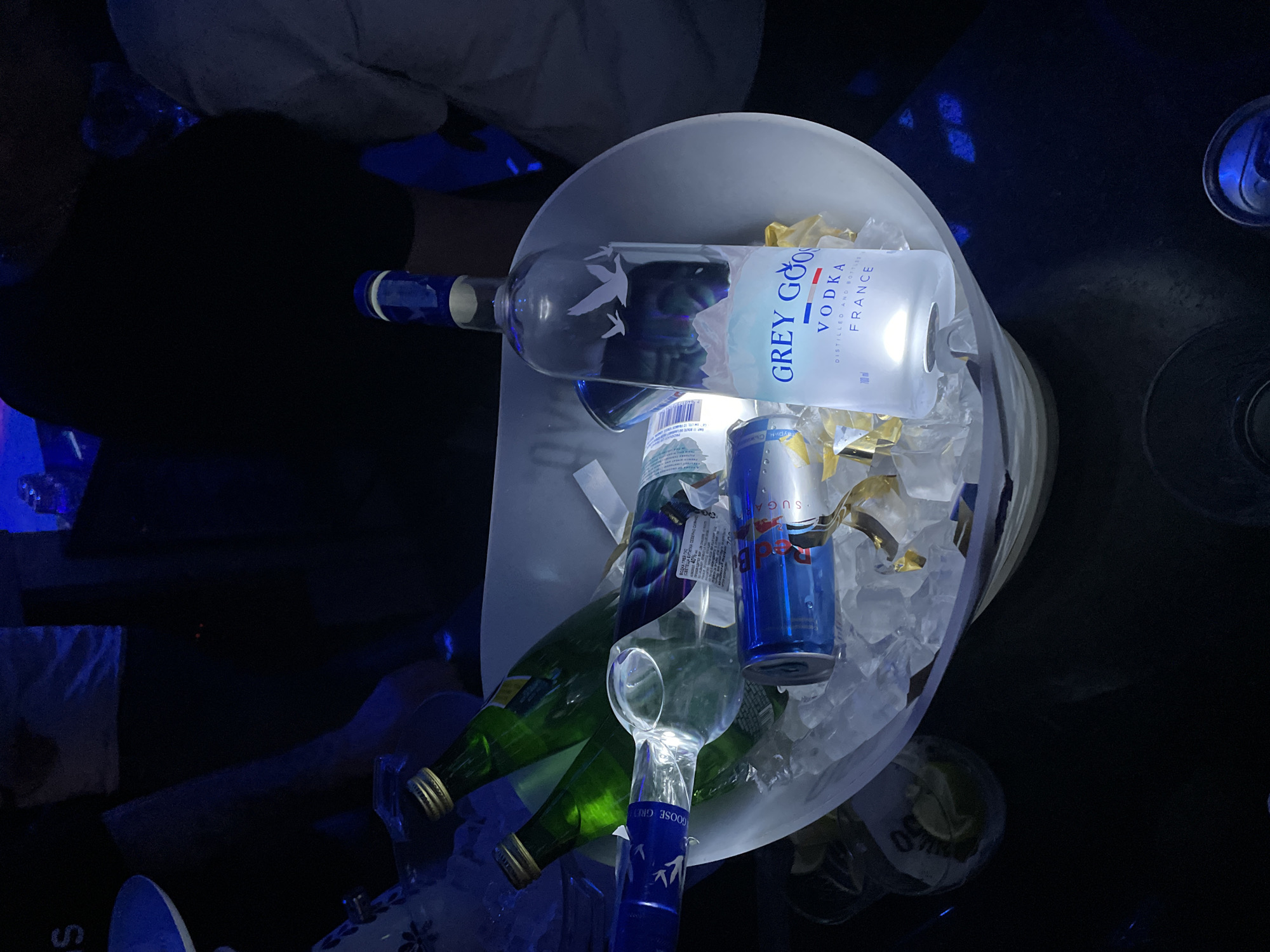 A white ice bucket containing a bottle of Grey Goose vodka, two green bottles (likely sparkling water or tonic), a can of Red Bull, ice cubes, and gold confetti is at the center of the image. The bucket is positioned on a dark surface that appears to be a table or countertop. In the background, there is a person partially visible. The lighting is dark with a blue tint, suggesting a bar or club setting. While the image cannot determine if the location is in Plovdiv, Bulgaria, the setting suggests a social gathering or celebration in a commercial entertainment venue.