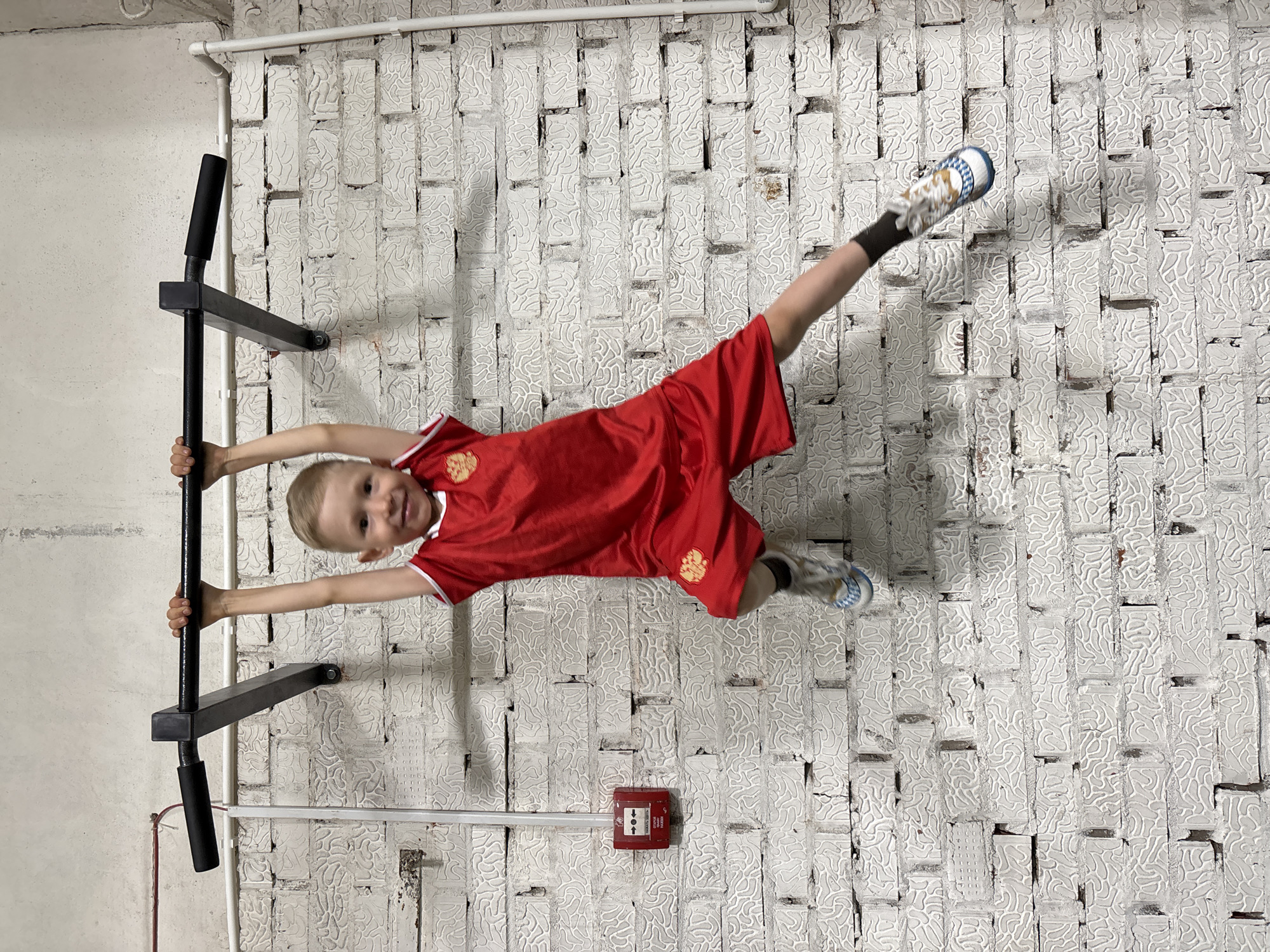 A young boy, clad in a red sports uniform with gold detailing, is hanging horizontally from a black, wall-mounted pull-up bar. His body is parallel to the white brick wall, with his hands gripping the bar and his legs extended to the right. He is wearing socks and athletic shoes.
The wall behind him is composed of white-painted bricks with a textured surface. A white pipe runs horizontally along the wall near the top of the frame. Below the boy, a red fire alarm pull station is mounted on the brick wall.
The location is likely an indoor space, given the construction of the wall and the presence of the pull-up bar. Based on the uniform and the prompt, this scene is likely located in Chaikovsky, Russia.