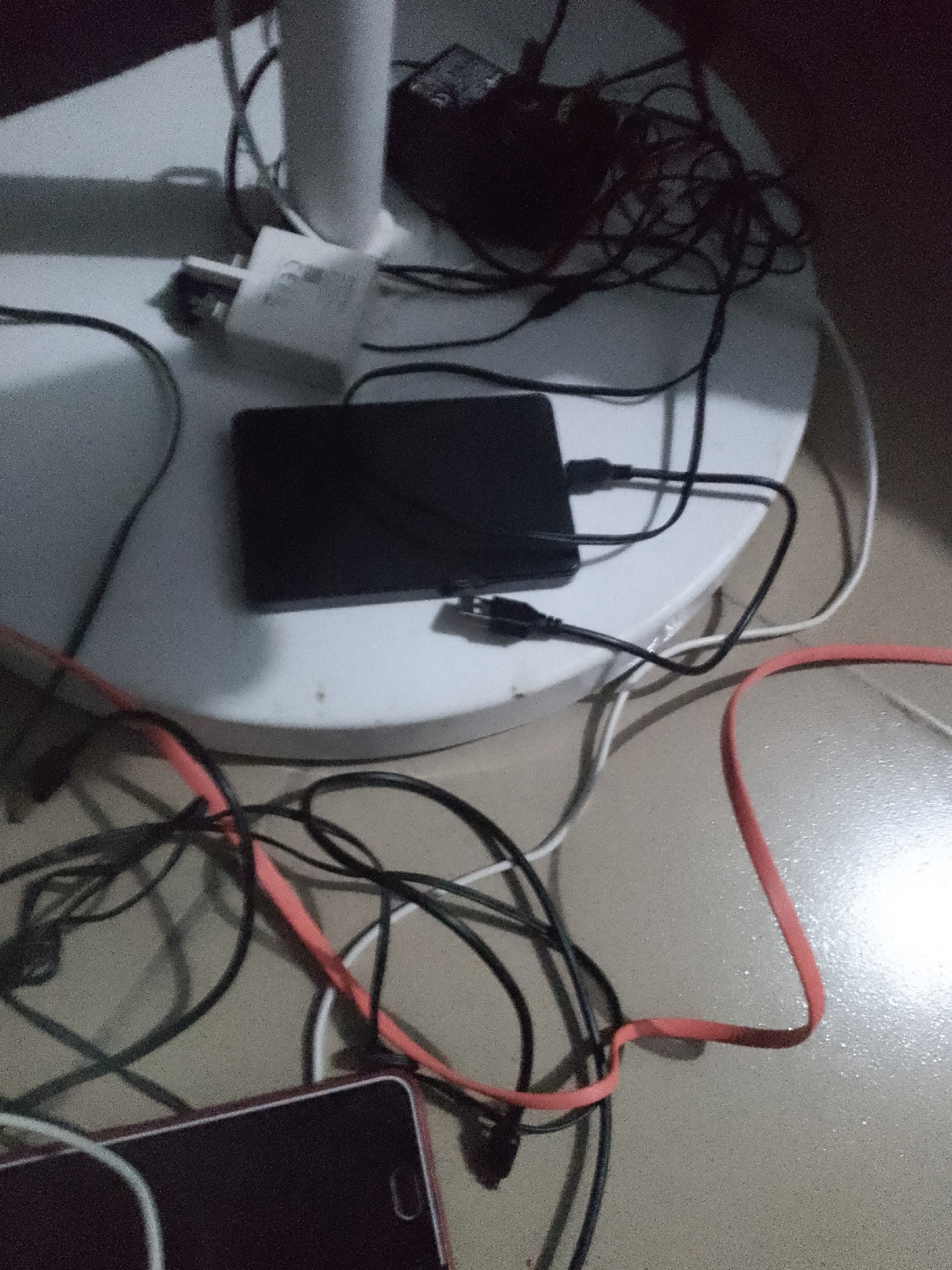 A black external hard drive is connected to cables and sits on a white surface. The surface appears to be the base of a fan. A white power adapter is also on the surface. Several black and orange cables are scattered around the hard drive and the base of the fan, with some extending out of frame. A portion of a dark-colored smartphone is visible in the foreground, also with cables attached. The background is dark and out of focus. The scene appears to be an indoor environment, possibly a desk or floor area.
