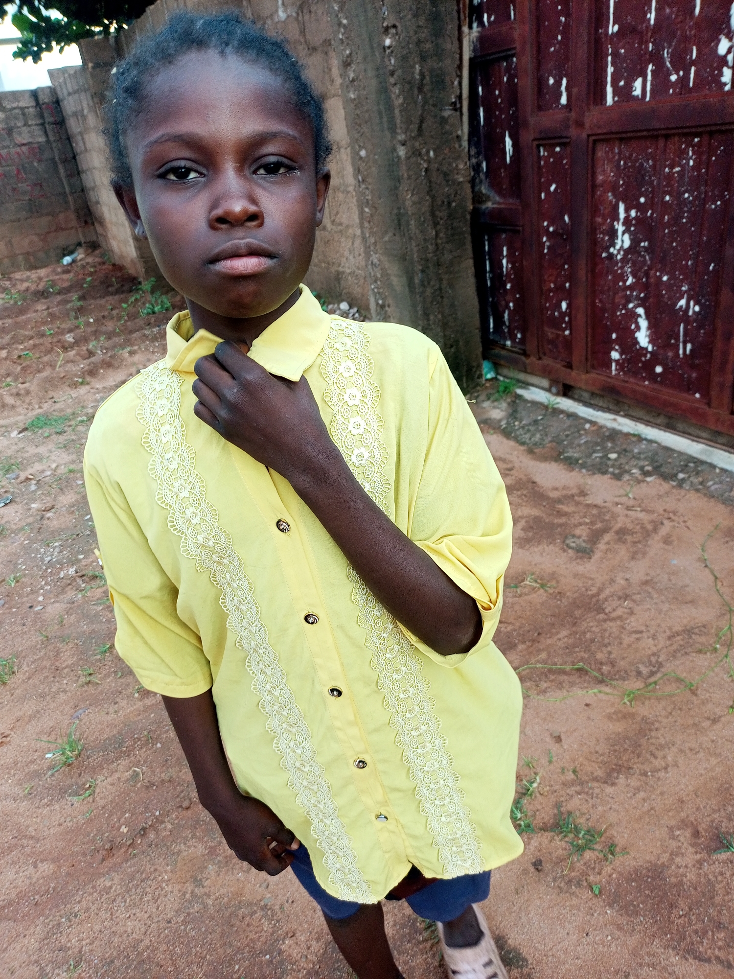 A young girl with dark skin and hair is standing in an outdoor, possibly rural, setting. She is looking directly at the camera with a neutral expression. She is wearing a bright yellow button-down shirt adorned with white lace trim down the front. The shirt has its collar popped, and she is holding one hand up to her neck, near the collar. She is also wearing blue shorts and light-colored sandals.
The background consists of a reddish-brown dirt ground, some sparse green vegetation, a rough-textured wall made of concrete blocks to the left, and a large, weathered, reddish-brown metal door or gate to the right. A tree trunk is visible between the girl and the door. The lighting suggests it is daytime, possibly late afternoon due to the warm tones. The overall scene appears to be in Gombe, Nigeria, based on the provided location context. There is no visible text in the image.