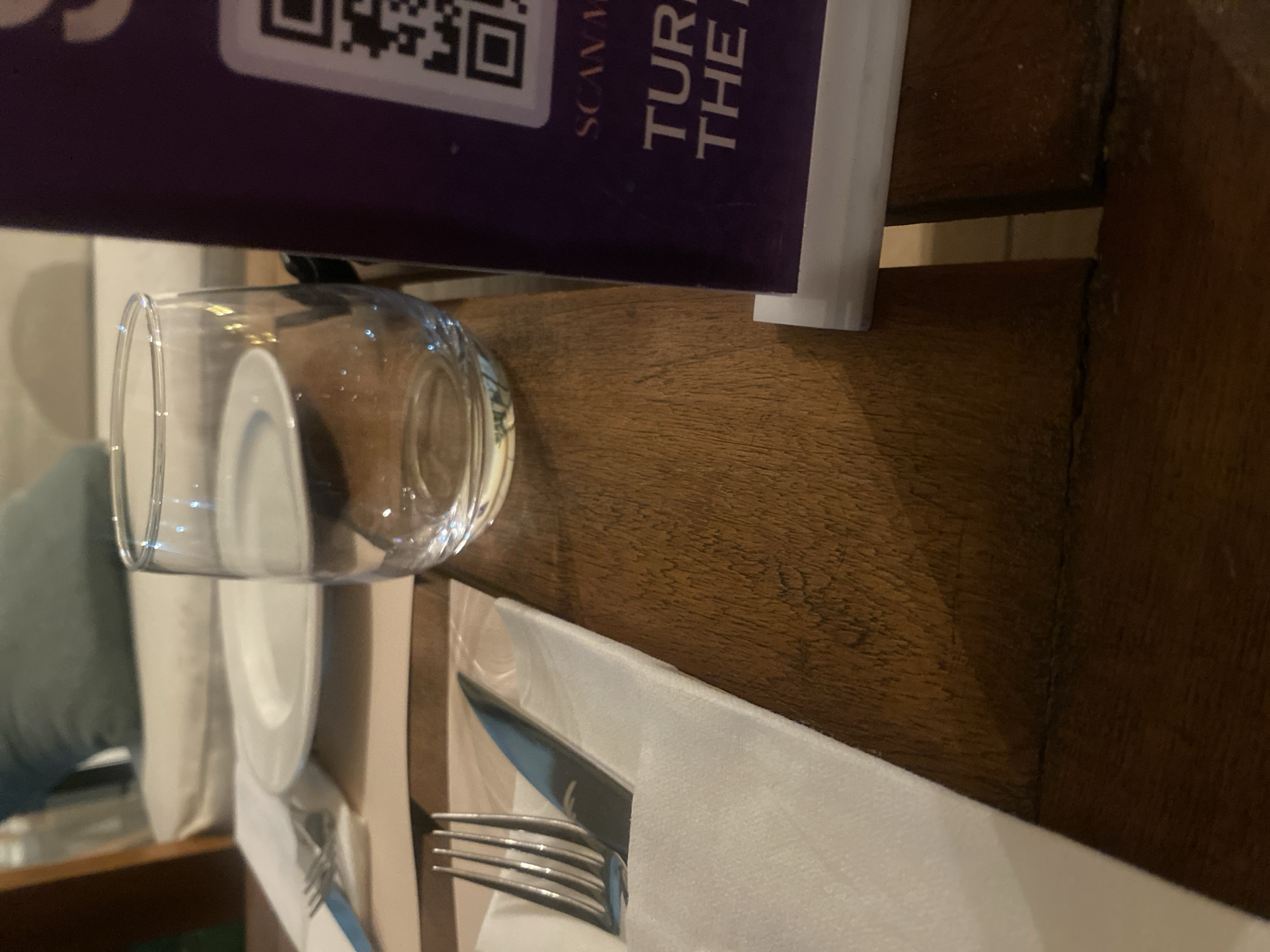 This image shows a close-up view of a table setting with a glass of water, silverware, and a menu. The subjects are primarily objects arranged on a wooden table. The scene appears to be indoors, possibly in a restaurant or dining area.
The activity taking place is a meal service or preparation for one. A clear glass is filled with water, and a fork and knife are neatly placed on a white napkin, suggesting readiness for dining. The menu, partially visible, adds to the context of a place where food is served.
Notable details include the texture of the wooden table, which appears to be rustic, and the clean, modern presentation of the tableware. The lighting is soft, hinting at an indoor environment, and the time of day is not explicitly discernible. The visible text on the menu is "SCANS" and "THE R," suggesting it's a promotional or informational item. The overall impression is one of organized hospitality.
The location context provided is Fagge A, Nigeria. While this information is external to the visual content of the image, it helps to contextualize the setting as likely a dining establishment within that region.