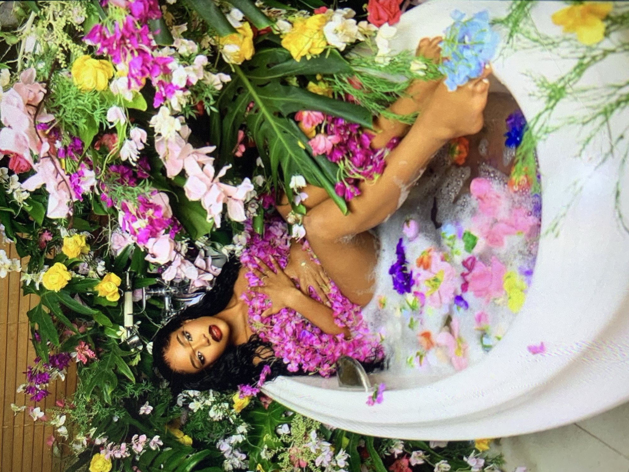 A woman reclines in a white bathtub filled with bubbly water and strewn with colorful flowers, amidst a dense arrangement of flowers and green foliage. The environment appears to be indoors, with a bamboo-like screen partially visible in the background. The woman's face is visible, and she appears to be looking at the camera. The flowers include pink, purple, yellow, and white varieties. The tub is surrounded by the foliage. The location is New Stadium, Nigeria.