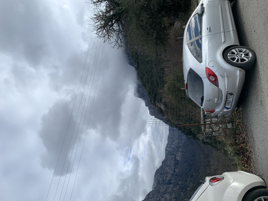 The image captures a rugged mountainous landscape in Vikos, Greece, under a heavily overcast sky. A narrow road in the foreground hosts two parked cars.
On the right side of the frame, a silver hatchback, identifiable as an Opel Corsa by its rear design, is parked facing away from the viewer. Its Greek license plate, "IHM 5799," is clearly visible. A white car's rear section is partially visible in the lower left foreground, also parked along the road.
The setting is characterized by steep, rocky mountains that rise majestically, their upper sections frequently obscured by dense, low-hanging grey clouds and mist, suggesting a high altitude or an approaching weather front. Sparse, bare-branched trees and dark, dense vegetation cling to the hillsides immediately behind the road. A low stone wall and a metal mesh fence line the edge of the road, offering a glimpse into a deep valley or gorge beyond. Several thin electrical or telephone wires stretch horizontally across the mid-ground, cutting across the cloudy sky.
The scene depicts a quiet moment along a mountain route, with the cars stationary, possibly at a scenic viewpoint. The time of day appears to be daytime, but the heavy cloud cover diffuses the light, creating a muted and somewhat somber atmosphere. No people are visible, emphasizing the raw, untamed nature of the landscape. The weather is distinctly cloudy and cool, with the mist adding to the dramatic mountain scenery.