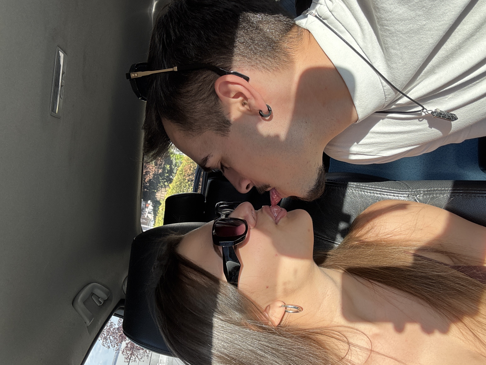 A photograph depicts two individuals kissing inside a car. The male has dark hair, a trimmed beard, an earring in his left ear, and wears sunglasses pushed back on his head. He is wearing a white T-shirt and a necklace with a pendant. The female has brown hair, wears sunglasses and earrings. The interior of the car is visible, including the dashboard, seats, and a view outside the window showing greenery and buildings. The location in the city of Sofia, Bulgaria, is not discernible from the image.