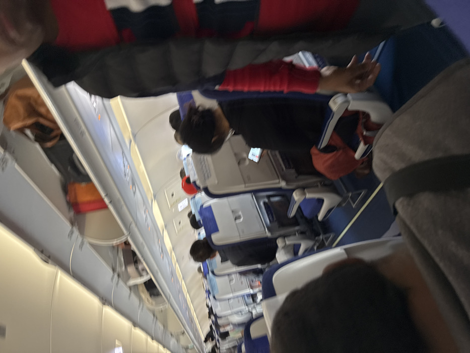 The image shows the interior of an airplane cabin during flight. Passengers are seated in rows of blue and white seats, some using tray tables. Overhead compartments are visible along the top of the cabin, with personal belongings stored inside. The overall lighting is dim. People can be seen seated in rows, including someone wearing red.