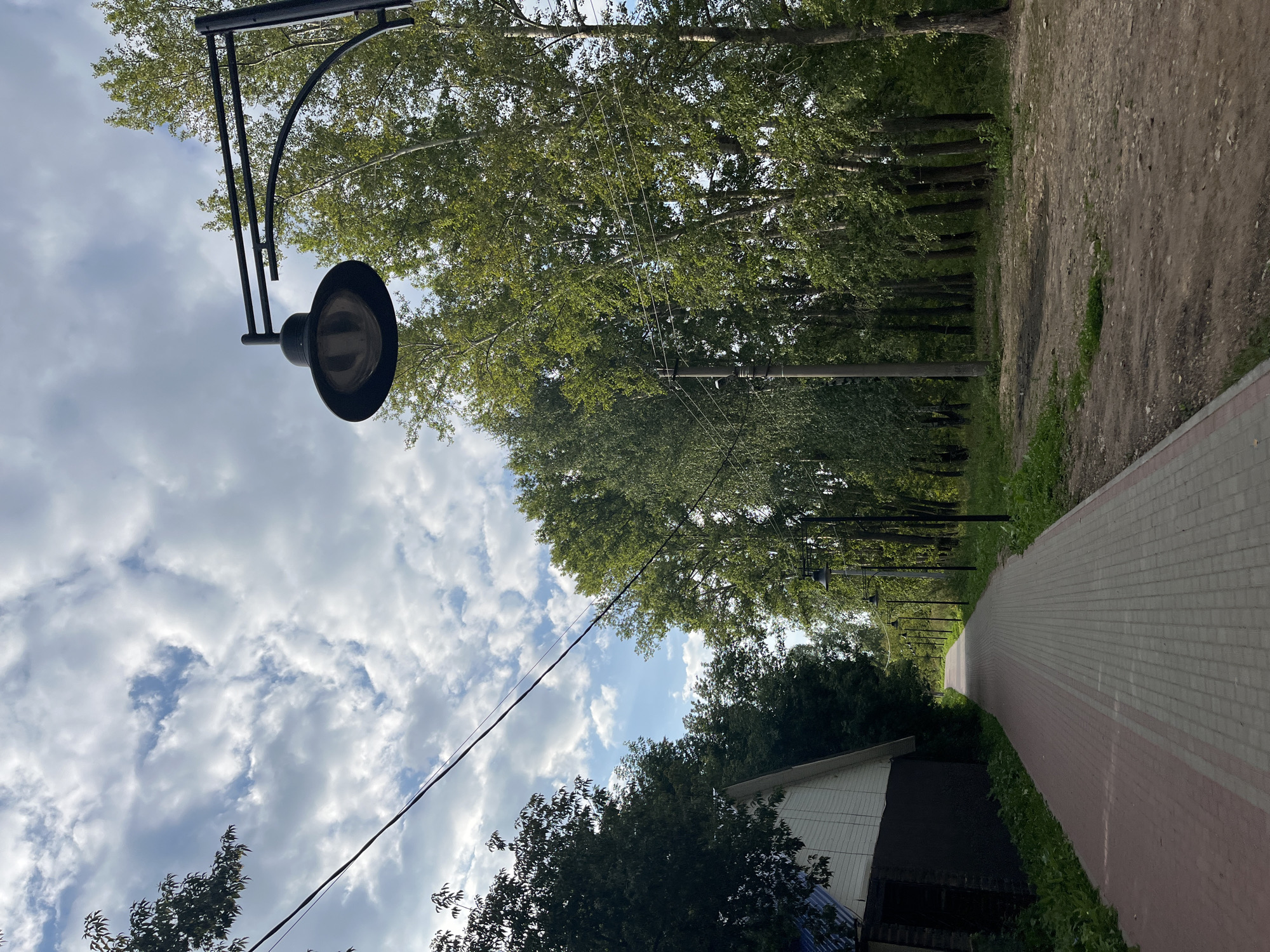 The image depicts a paved pathway running alongside a wooded area and a building. The pathway is constructed with red and gray paving stones and is bordered by grass and dirt on the right side, and grass and a building on the left. A prominent black lamppost with a wide, bowl-shaped light fixture is positioned in the foreground on the right. Several similar lampposts are visible further down the pathway, receding into the distance. A line of trees, predominantly deciduous with green foliage, forms a dense barrier along the right side of the pathway. The building on the left has white siding and a dark-colored foundation or lower level, with a blue roof. Power lines are strung across the sky, intersecting with the trees and lampposts. The sky is partly cloudy, with a mix of white and gray clouds against a blue background. The overall scene suggests an outdoor public space, possibly a park or a suburban street with pedestrian access. There are no people or animals visible in the image. The lighting suggests it is daytime, likely late afternoon or early evening given the angle of the shadows.