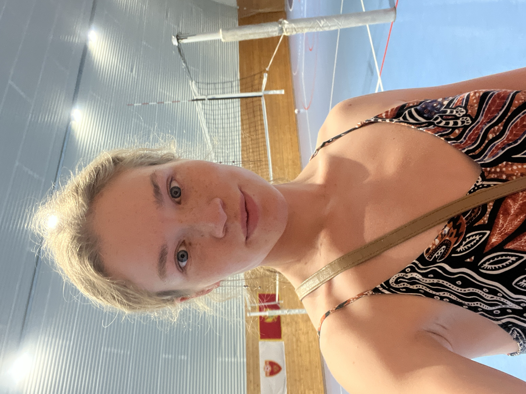 A fair-skinned woman with blonde hair and blue eyes is posing in an indoor sports venue. The background shows a volleyball net and court with light blue flooring and wooden paneled walls. Two flags are visible on the wall. The woman is wearing a printed top. The location is likely a sports hall in Pobrdje, Montenegro.