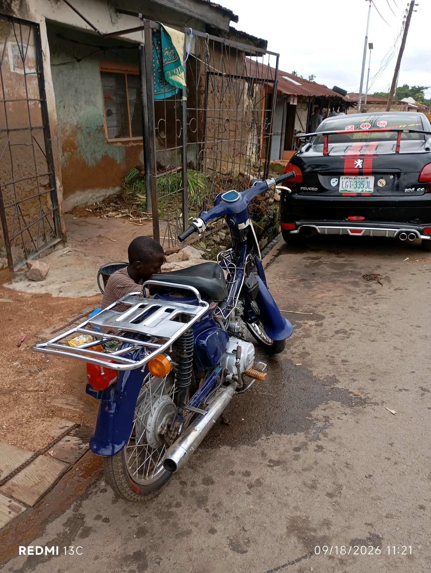 A young boy wearing a striped shirt is partially visible, kneeling next to a blue motorcycle parked on an unpaved road. The motorcycle has a chrome luggage rack. In the background, a black Peugeot 206 is parked. Behind the vehicles are dilapidated buildings with corrugated iron roofs, suggesting a rural or semi-urban environment. A piece of patterned fabric is hanging on a metal structure to the left of the buildings. The ground is a mix of dirt and asphalt, with some wet patches and debris. The scene is likely in Shendam, Nigeria, as indicated by the timestamp and phone model.