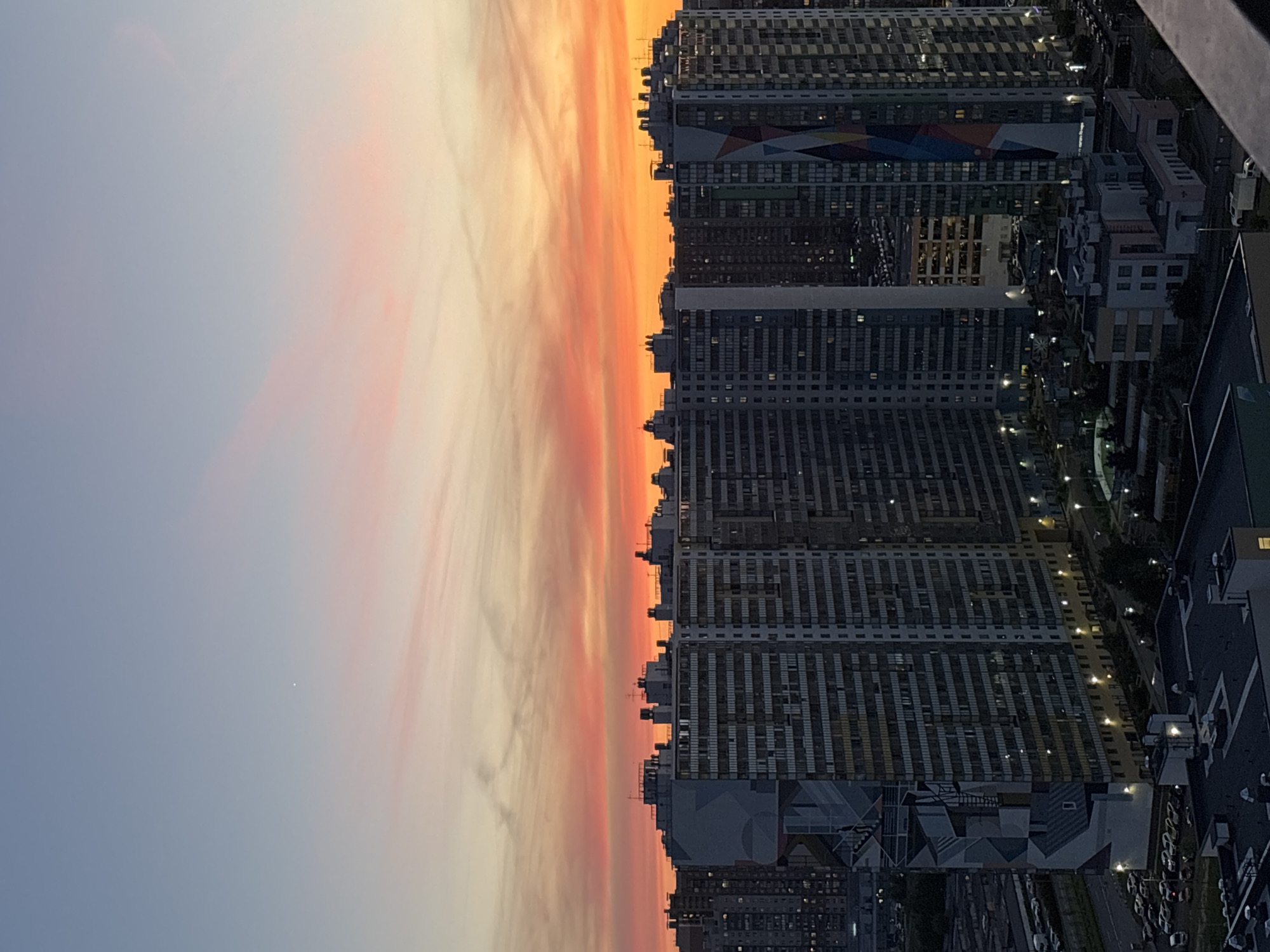 A high-angle, medium shot captures a city skyline at dusk. The sky displays a gradient of soft blues at the top, transitioning into vibrant hues of pink, orange, and yellow near the horizon, indicative of a sunset. A single bright star or planet is visible in the upper left quadrant of the sky. Dominating the lower portion of the frame are several tall, modern apartment buildings. These structures are characterized by their repetitive window patterns and are illuminated by artificial lights. One building on the left features a large, abstract geometric mural in shades of grey, white, and blue. Another building on the right has a colorful, angular mural composed of red, blue, yellow, and white. The foreground shows the tops of lower buildings and what appears to be a rooftop. In the mid-ground, streetlights illuminate parking lots and roads where cars are parked. The overall scene depicts a residential urban environment during the transition from day to night.