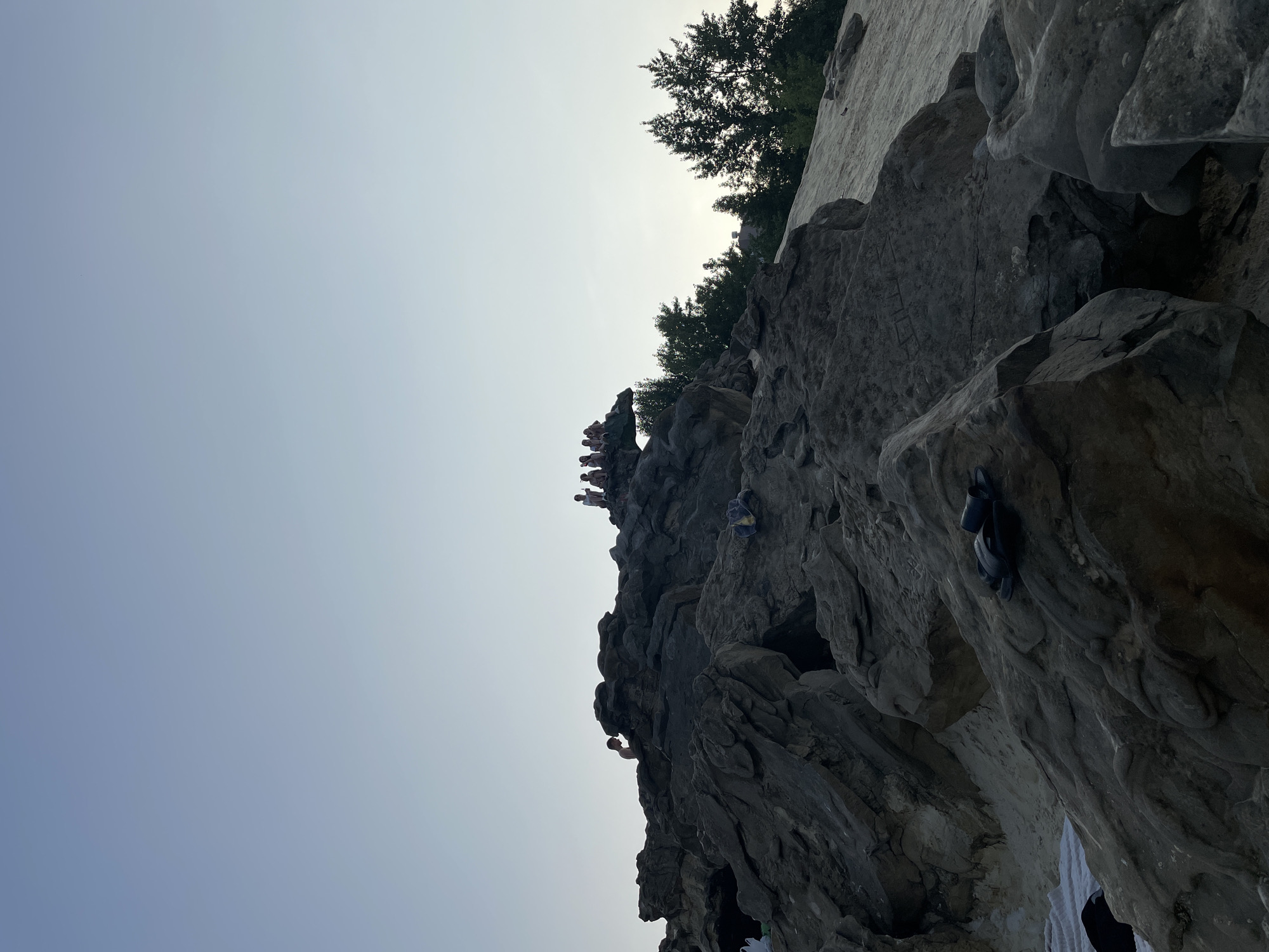 The image depicts a natural scene, likely a rocky area with sand, under a clear sky. In the foreground, large, rough, gray rocks dominate the view, with a pair of black flip-flops resting on one of the rocks. A patch of white fabric, possibly a towel, is also visible near the bottom left. Further into the scene, a group of people, silhouetted against the sky, are seated on top of a rocky outcrop. One individual is standing and looking down from a higher rock face. In the background, several trees with dark green foliage are visible, and a faint suggestion of a building can be discerned between them. The overall lighting suggests it might be late afternoon or early evening. This scene is not specifically identifiable as being located in the city of Tambov, Russia, based on the visual information provided.