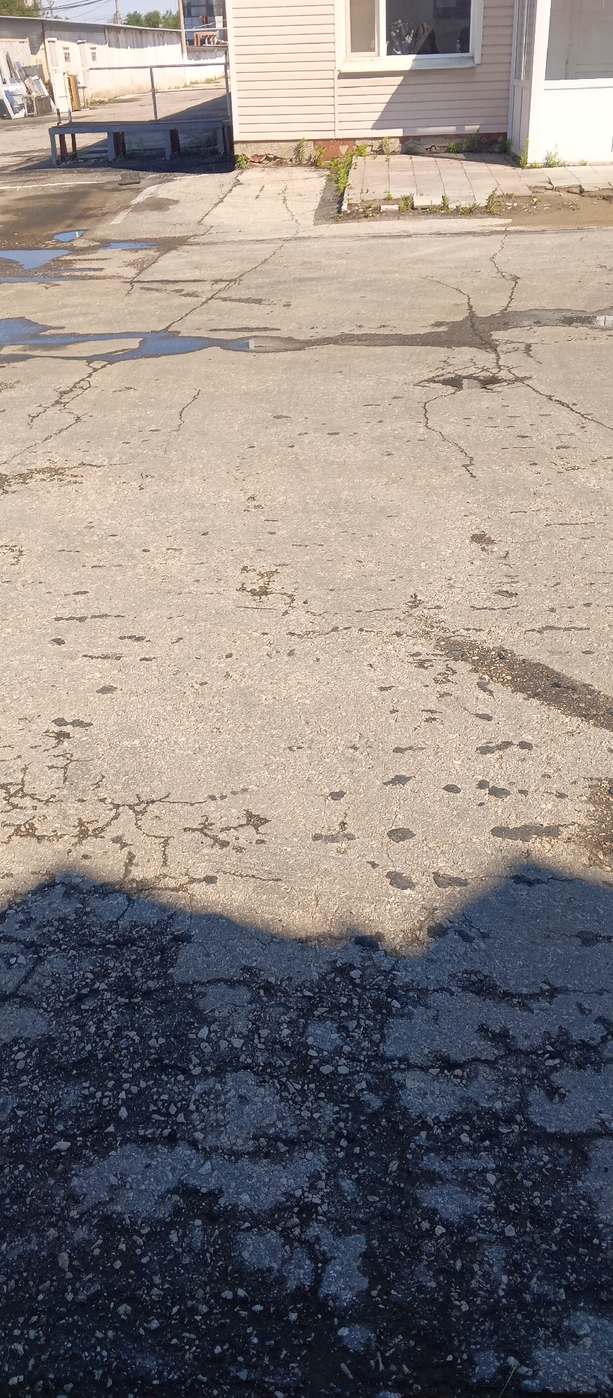 The image shows a cracked asphalt surface in front of a light-colored building. The building has horizontal siding and a window. There is a concrete pathway in front of the building, and patches of grass are growing through the cracks. The asphalt surface is wet in several places, and there are dark stains that appear to be oil or grease. In the foreground, there is a large shadow cast across the asphalt. The background shows more of the building and some industrial-looking structures. The overall scene suggests an outdoor area of a commercial or industrial property.
