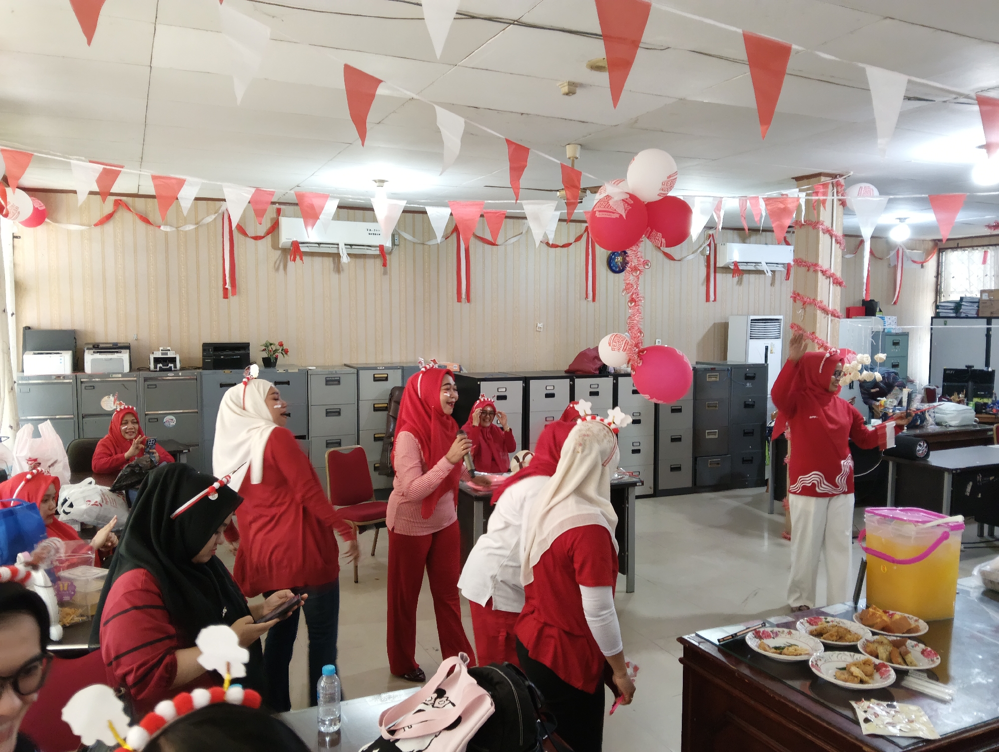 The image depicts an indoor gathering, likely an office party, decorated with red and white triangular banners and balloons. Several individuals, mostly women wearing headscarves and red or white attire, are present. Some are standing, interacting, and appear to be celebrating, while others are seated. There are also office furniture such as filing cabinets, printers, and desks visible in the background. A table in the foreground displays plates of food and a large jug of orange liquid. The overall atmosphere suggests a festive occasion, possibly related to Indonesian Independence Day given the prominent red and white theme. The image is taken from eye-level. It is impossible to determine the specific location in Pekanbaru, Indonesia, without additional contextual information.