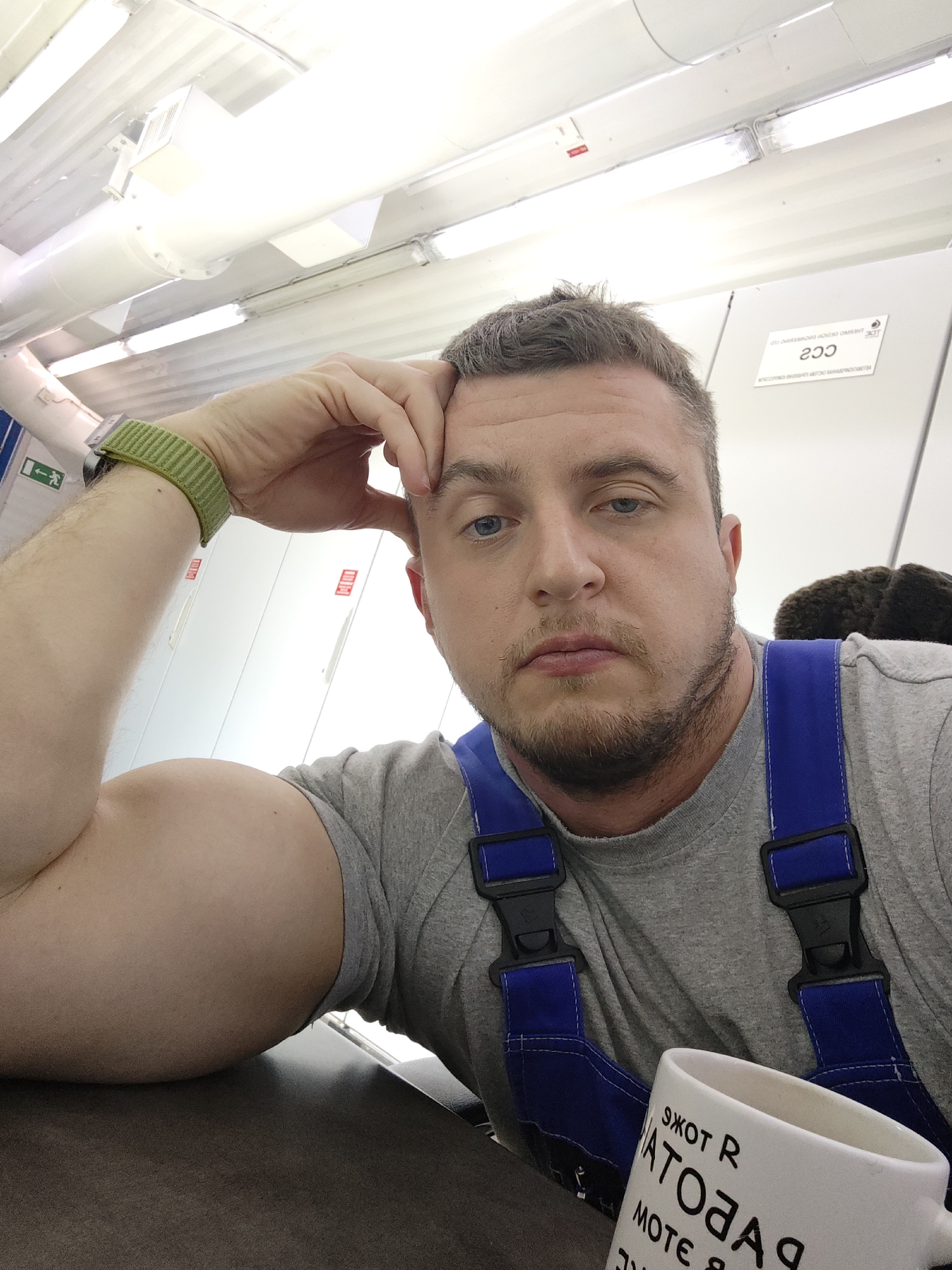 A man with a slightly disheveled grey haircut, light blue eyes, and a stubbly beard is shown in a close-up, selfie-style photograph. He is wearing a grey t-shirt with blue overalls. His left arm, showing a green smartwatch band, is resting on a surface, with his hand supporting his forehead. A white mug with text in a non-English language is visible near the bottom right of the frame. The background shows a white interior, likely an industrial or commercial setting, with overhead lighting fixtures. A white panel with the marking "CC2" is visible in the background. The location is listed as Unknown, Unknown.