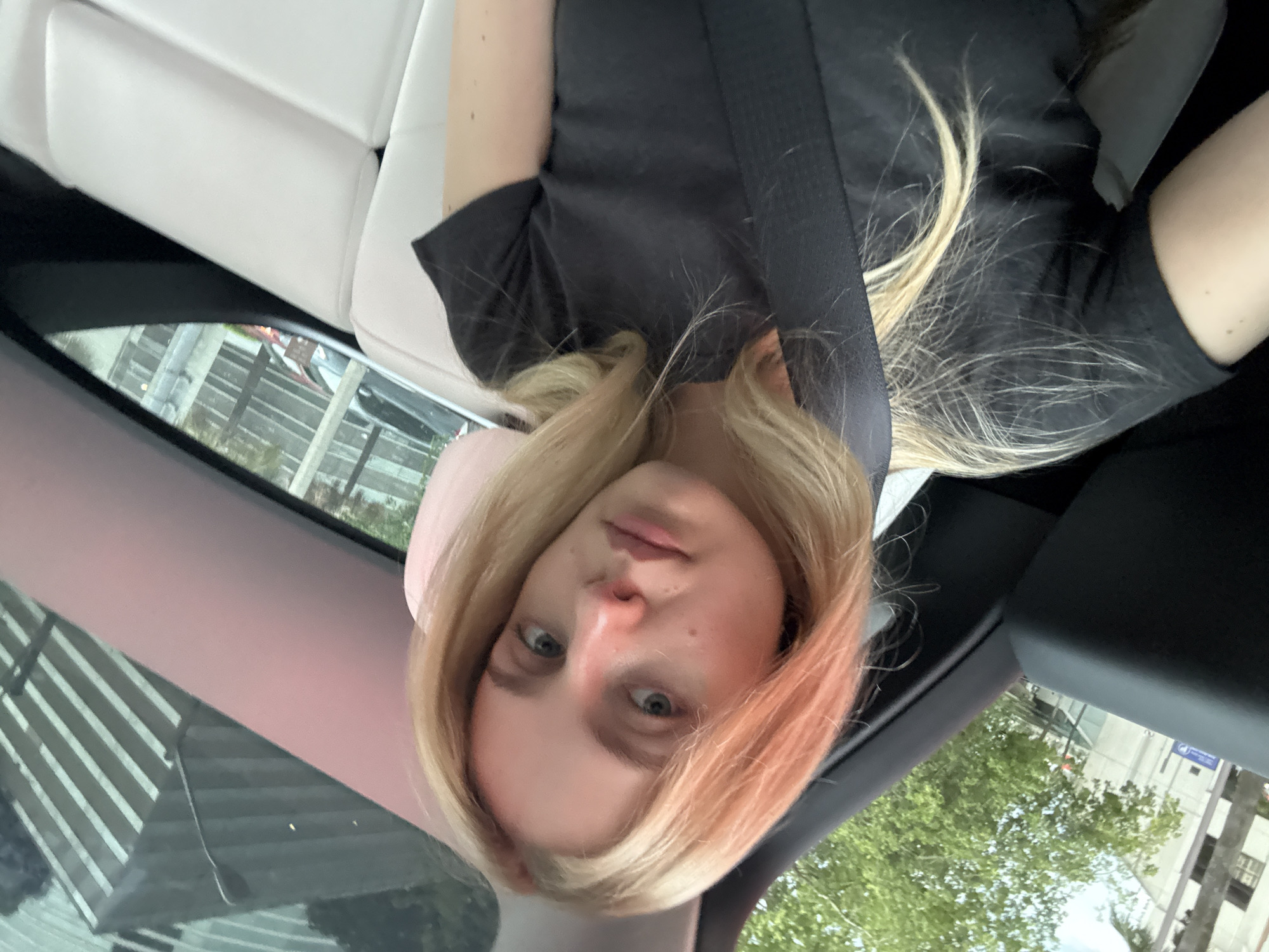 A young woman with blonde hair is seated in the back of a car. She is wearing a dark t-shirt and a seatbelt. The interior of the car appears to be white. Through the windows, a city environment is visible, with buildings and trees. The lighting suggests it is daytime.
