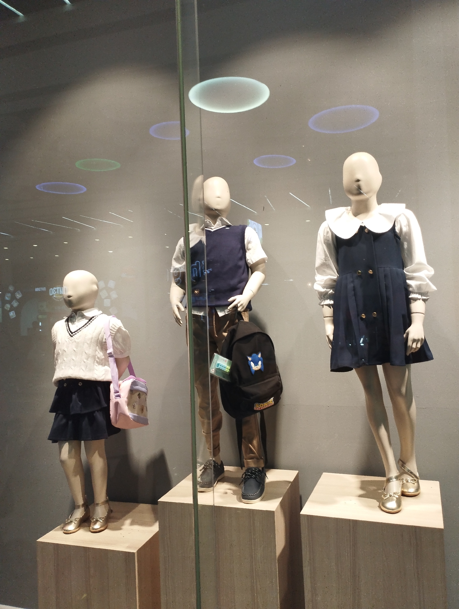The image shows three child mannequins in a storefront window display. The mannequin on the left wears a white cable-knit sweater vest over a white collared shirt, a navy skirt, and gold Mary Jane shoes. It also has a pink and purple backpack slung over its shoulder. The middle mannequin wears a navy vest over a white collared shirt and tan pants, with black lace-up shoes and a black backpack. The mannequin on the right wears a white collared shirt with a navy pinafore dress and gold ballet flats. The display is set against a grey wall with reflections of circular lights on the ceiling. This scene is from a storefront in the city of Dushanbe, Tajikistan, as indicated by the text "OSTHUD" reflected on the window, which is an abbreviation for "Ostoni Hudud," meaning "Border Region" in Tajik, a known retailer in Dushanbe.