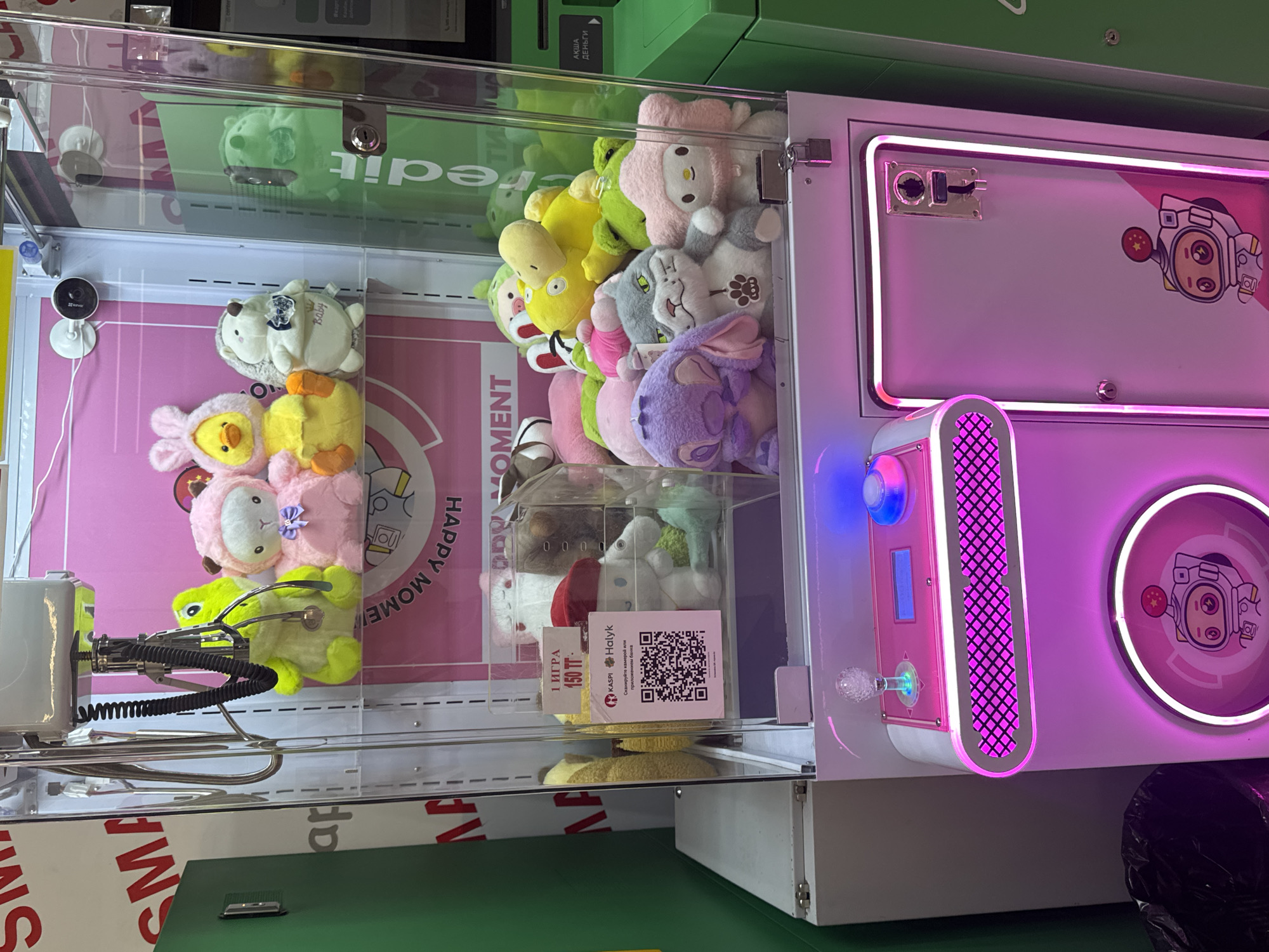 The image shows the interior of a crane game machine filled with plush toys, located in Kokshetau, Kazakhstan. The machine is pink and white with neon purple lighting around the bottom compartment. The crane mechanism is visible at the top. Plush toys include a pink sheep, a yellow duck, a hedgehog, a gray cat, a purple character resembling Stitch, and a pink bear. A QR code for payment is visible on a red and white sign, alongside text in Russian indicating "1 IGRA 150 TT" (1 game 150 Tenge). The background features a pink wall with "HAPPY MOMENT" printed on it. A green vending machine is partially visible to the right of the crane game.