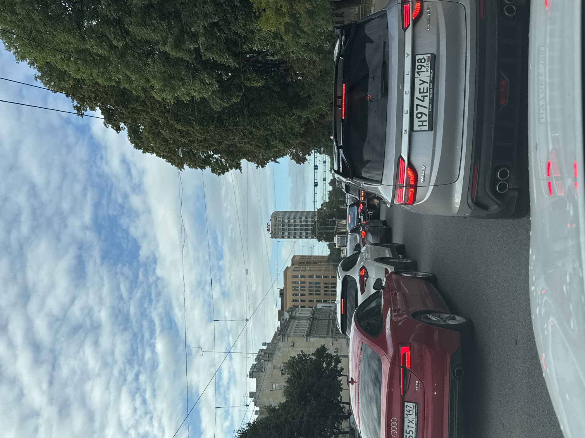 The image captures a traffic jam on a city street, likely in Saint Petersburg, Russia, given the license plate format and a visible "BCC" building which is a known landmark in the city. The perspective is from inside a vehicle, looking forward. In the foreground, the rear of a silver Geely Coolray SUV is visible, displaying the license plate H974EY 198. To the left, a red Audi A5 coupe is also in traffic, with license plate 655TX 147. The street is lined with trees on the right and buildings on the left. In the background, there are several multi-story buildings, including a taller, slender building with a distinct architectural style, and a distinct tower with "BCC" signage. The sky is partly cloudy. The traffic appears to be at a standstill.