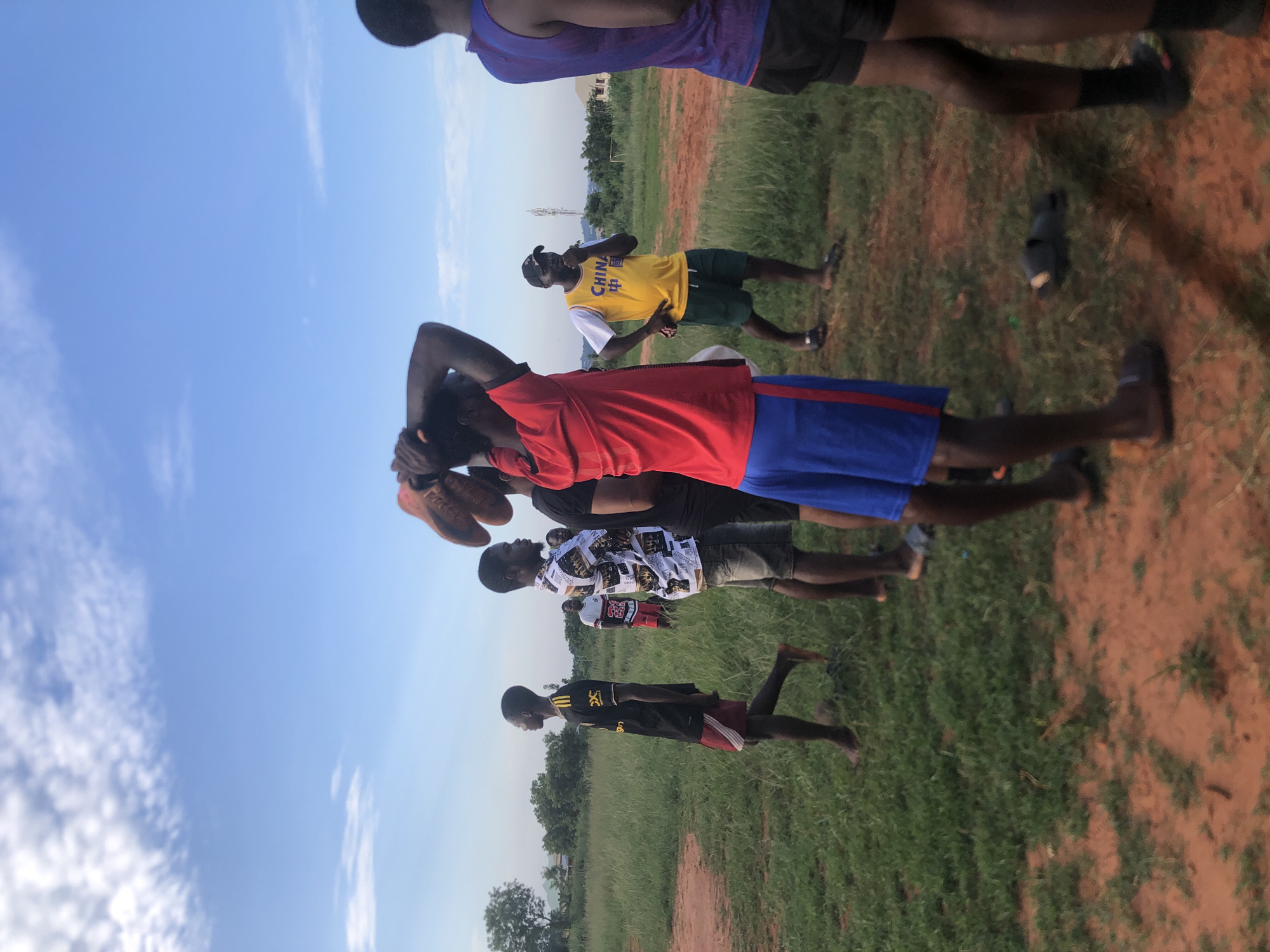This image captures a group of young men and boys gathered in an open, grassy field under a clear blue sky with scattered white clouds. The setting appears to be late afternoon or early evening, characterized by bright, natural light. The ground consists of green grass interspersed with reddish-brown dirt paths.
In the foreground, a young man wearing a red short-sleeved shirt and blue shorts is holding a pair of brown sports shoes or cleats above his head, possibly engaging in a stretch or preparing for an activity. To his right, another man stands prominently in a yellow sleeveless shirt, which features the word "CHINA" in black letters and Chinese characters (likely "中国") beneath it. He is also wearing dark shorts and a baseball cap, and he looks towards the viewer with a casual hand gesture.
Behind these two, a man in a black and white patterned shirt and dark shorts is visible. Further left, a young boy in a black and red shirt with a partially visible logo (resembling Adidas stripes) and red shorts stands barefoot. Several other individuals are partially visible in the background and foreground, indicating a larger gathering. Sandals and other footwear are scattered on the ground near some participants. In the distance, low-rise buildings and trees suggest a semi-rural landscape. The overall atmosphere is casual, indicative of a sports practice, a game, or a social gathering in Nsugbe, Nigeria.
