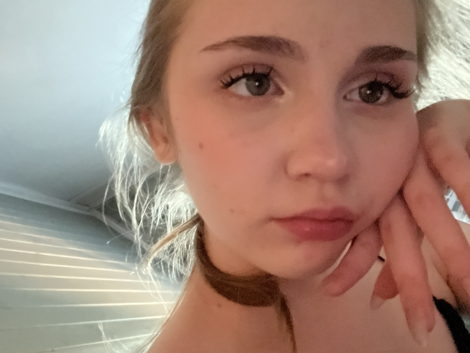 The image is a close-up, low-angle, indoor, portrait of a young Caucasian female. She is looking at the camera, with her head tilted and resting on her hand. Her eyes are open and her lips are slightly parted, with a glossy pink lipstick applied. She has light brown hair, which is partially obscuring her face and neck. The background appears to be a room with vertical blinds casting shadows. There is no indication of the city of Ashkadar, Russia.