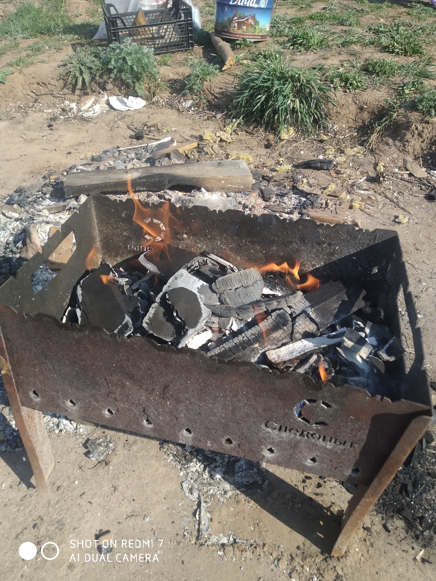 A close-up shot depicts a metal barbecue grill with burning charcoal. Flames rise from within the grill. The grill rests on a dirt surface. The words "Следопыты" are inscribed on the grill's side. The scene is outdoors, with grass and other vegetation visible in the background. The shot was taken with a Redmi 7 AI Dual Camera. The location appears to be Chaikovsky, Russia, based on the writing on the grill.