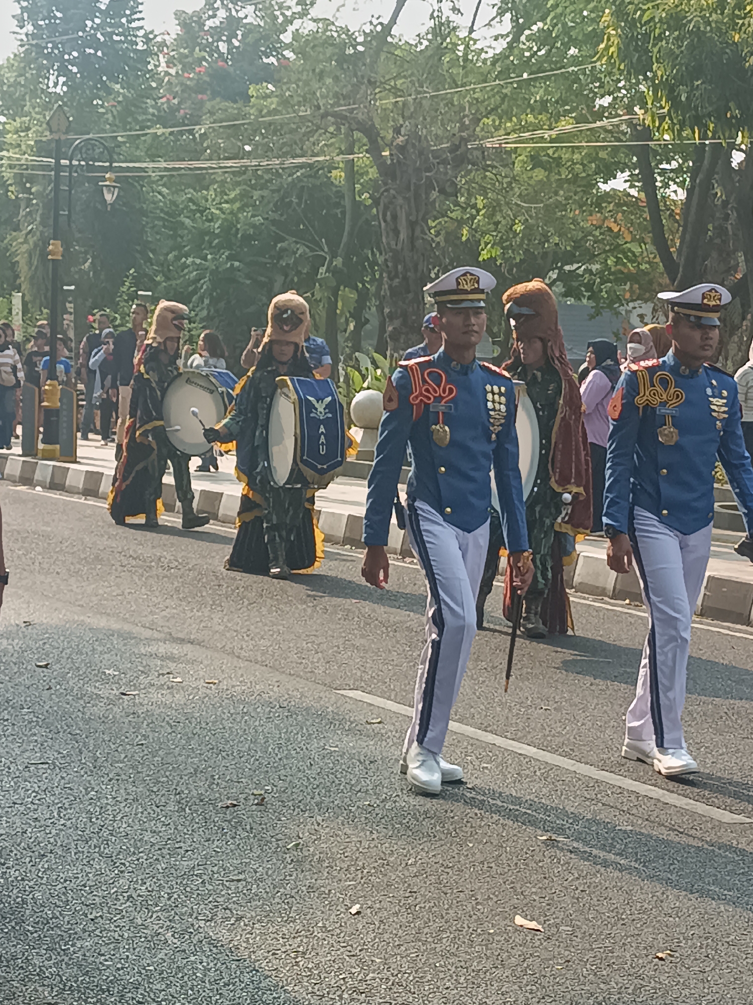 The image depicts a parade in Sleman, Indonesia. In the foreground, two young men in matching blue military-style uniforms with white pants and white shoes march in unison, holding batons. One of them has a dark baton. Behind them, several individuals are dressed in elaborate, traditional costumes adorned with animalistic headwear, carrying large drums. One of these individuals is beating a drum with a white drumstick. In the background, a crowd of spectators lines the street, some holding cameras. The scene takes place on a paved road with a sidewalk and a lamppost visible on the left. Lush green trees and foliage form a canopy overhead, suggesting an outdoor setting, possibly a park or tree-lined avenue. The lighting indicates it is daytime, with sunlight casting shadows on the road.