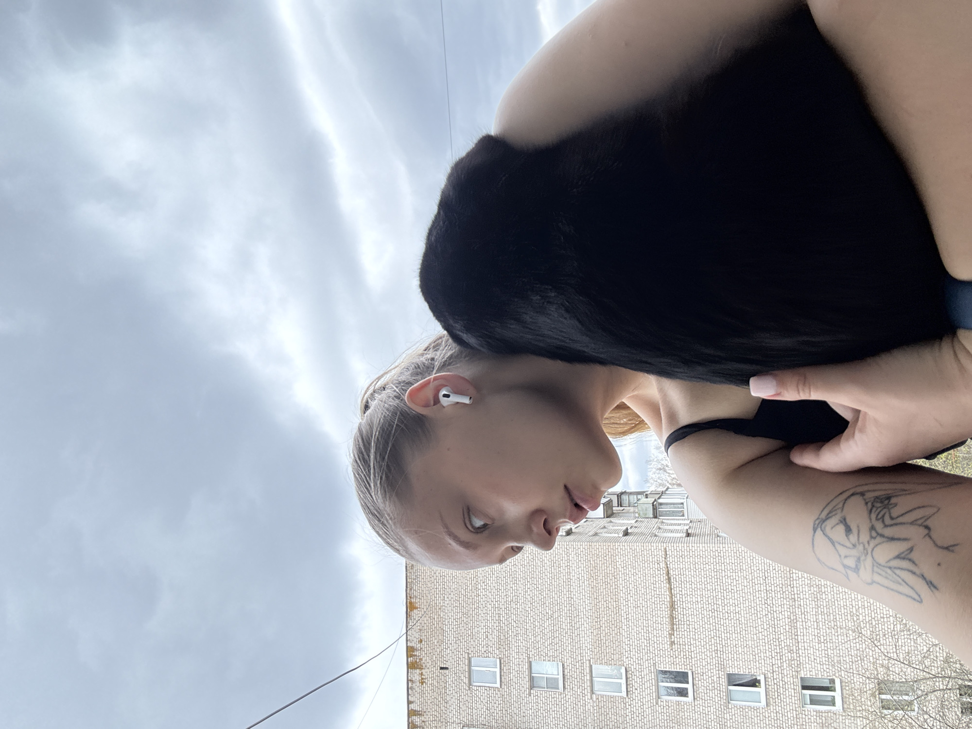 A young woman is depicted in a medium shot, oriented with the background building upright and the sky above. She has fair skin, light blonde hair tied back, and wears a black top with a visible strap. A white AirPods earbud is in her right ear. Her left arm features a distinct line-art tattoo, illustrating a stylized winged figure or mythical creature. Her left hand is resting near her bicep. She is holding or embracing a large, fluffy black object, likely a pet cat or small dog, which is pressed against her right shoulder and chest, obscuring its head. Her gaze is directed downwards and to her right, with a neutral expression and slightly parted lips.
The background consists of an overcast sky filled with light grey clouds, suggesting a cloudy day without direct sunlight. Below the sky, a large, light-colored brick building with multiple small, rectangular windows is visible, possibly residential or industrial architecture common in Chaikovsky, Russia. Bare tree branches are visible on the far right side of the image, indicating late winter or early spring. A thin black power line crosses the upper portion of the sky. No visible text is present in the image. The overall scene conveys a casual outdoor moment.
