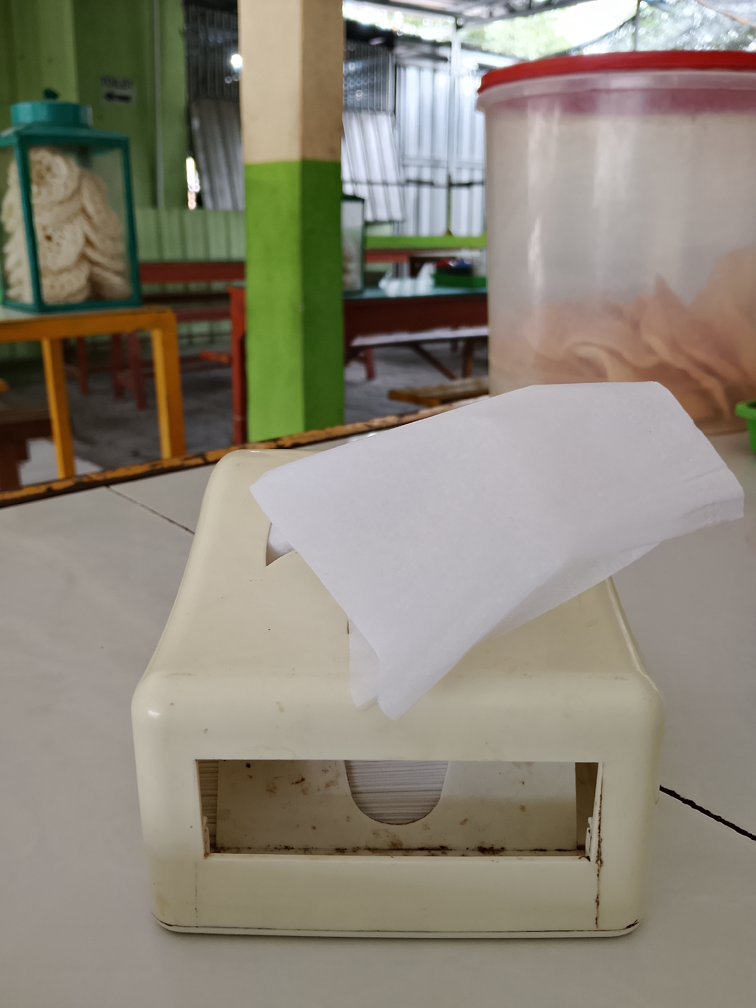 In Balun, Indonesia, a tissue box sits on a table in what appears to be a restaurant or food stall. The tissue box is cream-colored and shows signs of wear and tear, with some rust and dirt visible inside the opening. A few white tissues are sticking out of the top. In the background, there is a large clear plastic container with a red lid, filled with what looks like crackers or chips. Another container, possibly a lamp or display case, holds more crackers. The environment seems rustic, with a corrugated metal roof and wooden beams visible, suggesting an outdoor or semi-outdoor dining area. The overall impression is of a simple, casual eating establishment.