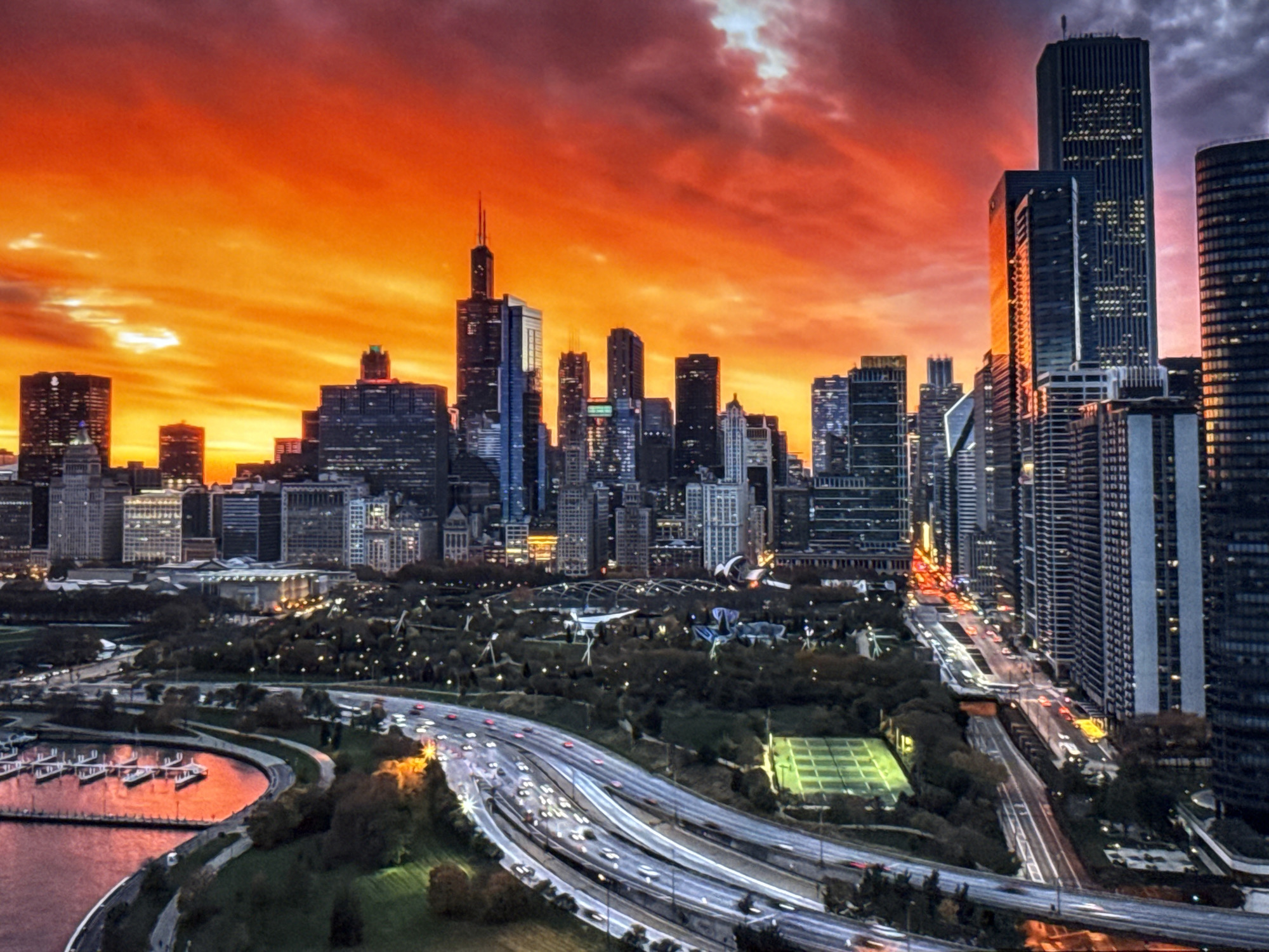 The image depicts a dramatic sunset over a city skyline. The sky is ablaze with intense hues of orange, red, and yellow, with some patches of bright white light and darker, purplish clouds. Silhouetted against this vibrant sky are numerous skyscrapers of varying heights and architectural styles. The buildings appear to be made of glass and steel, with many windows reflecting the fiery colors of the sunset.
In the foreground, a complex network of multi-lane highways can be seen, with streaks of light from vehicle headlights and taillights indicating movement. Trees and green spaces are visible between the highways and the cityscape, suggesting a park or urban green area. To the left, a body of water, possibly a lake or harbor, is present, with several boats docked in a marina. The water also reflects the intense colors of the sky, creating a mirror-like effect. The overall impression is one of a bustling urban environment bathed in the dramatic light of a sunset.
This image does not depict the city of Burgas, Bulgaria. The architecture and skyline are characteristic of a major North American city, likely Chicago, given the prominent skyscrapers that resemble iconic structures there.