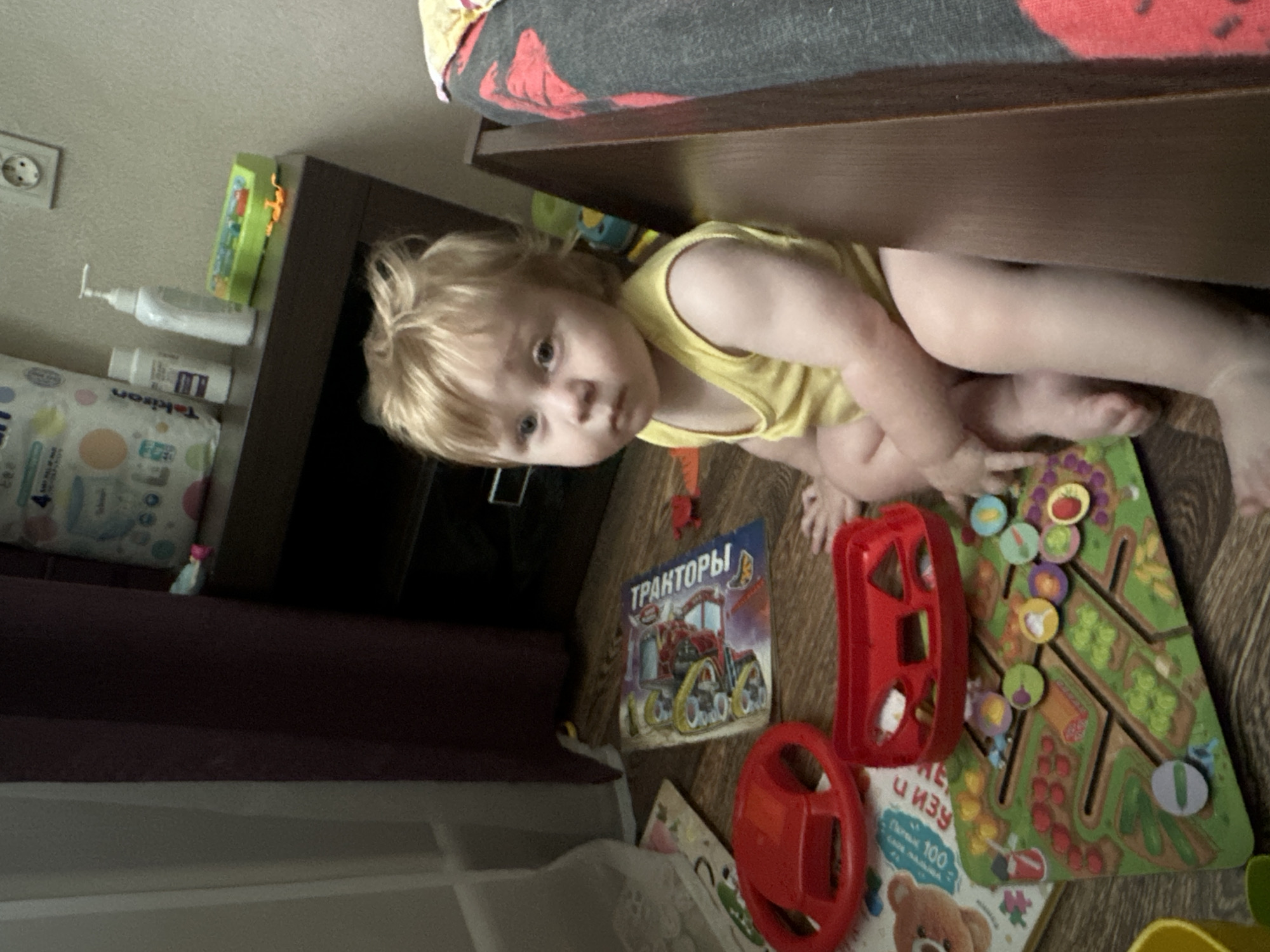 A fair-skinned toddler with blonde hair, wearing a yellow sleeveless shirt, sits on a wooden floor in a room, possibly a child's bedroom or play area. The toddler is looking forward with a neutral expression. Scattered around the toddler are several children's toys, including a red toy steering wheel, a red shape sorter, and a colorful play mat depicting a garden scene with fruits and vegetables. A children's book with images of tractors is also visible on the floor. In the background, a dark brown nightstand holds several items, including a package of baby wipes and bottles of lotion. The room appears to be lit by artificial light. The location is Sayanogorsk, Russia.