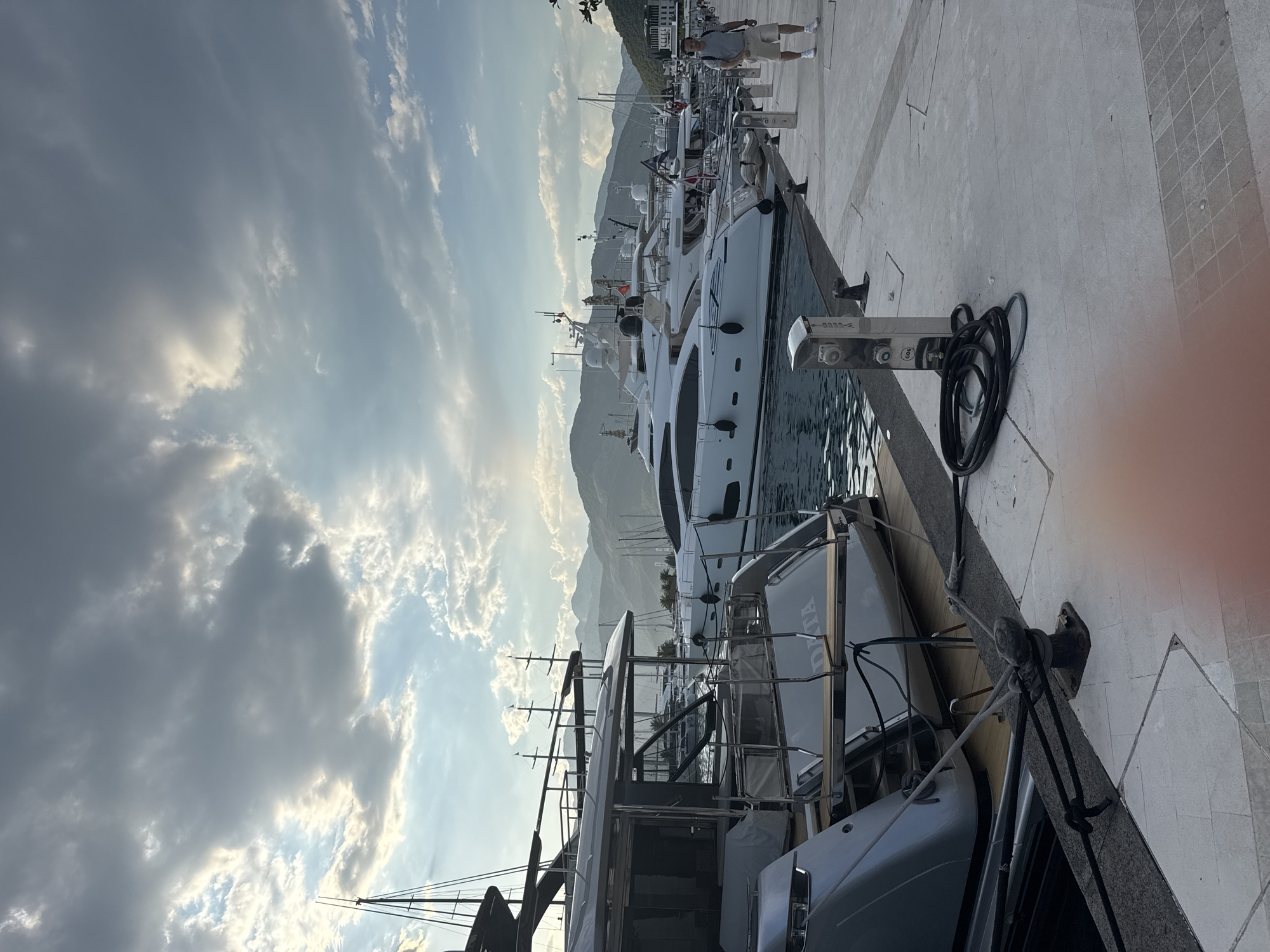 A man in shorts and a t-shirt walks along a paved pier in Tivat, Montenegro, past several docked yachts. The yachts are white and sleek, with dark trim and masts. The sky is cloudy with patches of sunlight. In the background, mountains rise behind the town. The scene appears to be in the Porto Montenegro marina, a luxury yachting hub in Tivat.