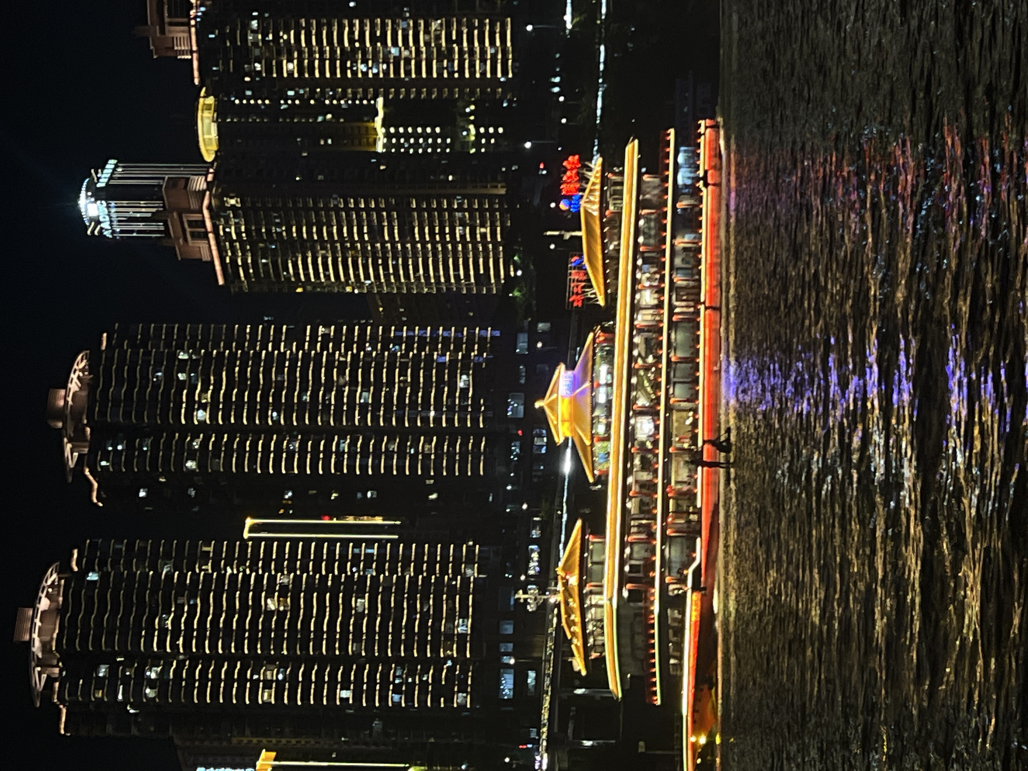 A night scene captured at 江北城街道, China, showcases a vibrant cityscape dominated by towering, illuminated skyscrapers that stretch into the dark sky. The buildings are adorned with numerous horizontal yellow lights, creating a striking pattern against their dark facades. In the foreground, a large, multi-decked boat, illuminated with red and yellow lights, cruises on a body of water. The boat features ornate, pagoda-like roofing structures in bright yellow and red. The water reflects the city's lights, creating a shimmering, colorful effect. Two figures are visible on the deck of the boat, and another two are standing near the water's edge on the boat's hull. The overall atmosphere is one of urban dynamism and nocturnal beauty.