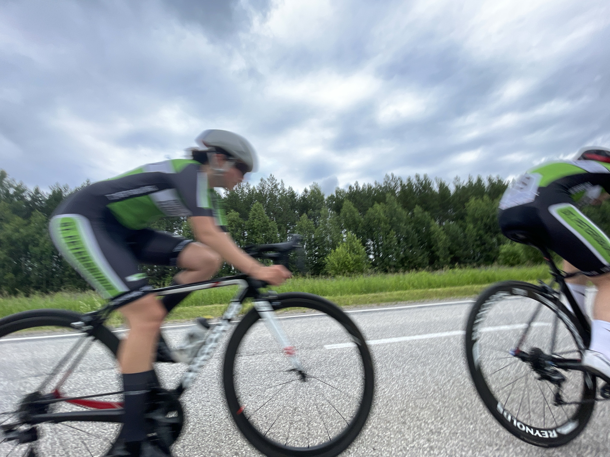 Two cyclists are racing on a road, with trees lining the right side of the road and a cloudy sky above. One cyclist is in the foreground, wearing a black and green cycling suit and a white helmet. Their bike is black with white lettering. The second cyclist is in the background, also wearing a black and green cycling suit and a helmet. The road has white lines marking the lanes. The image is blurry, suggesting motion.
