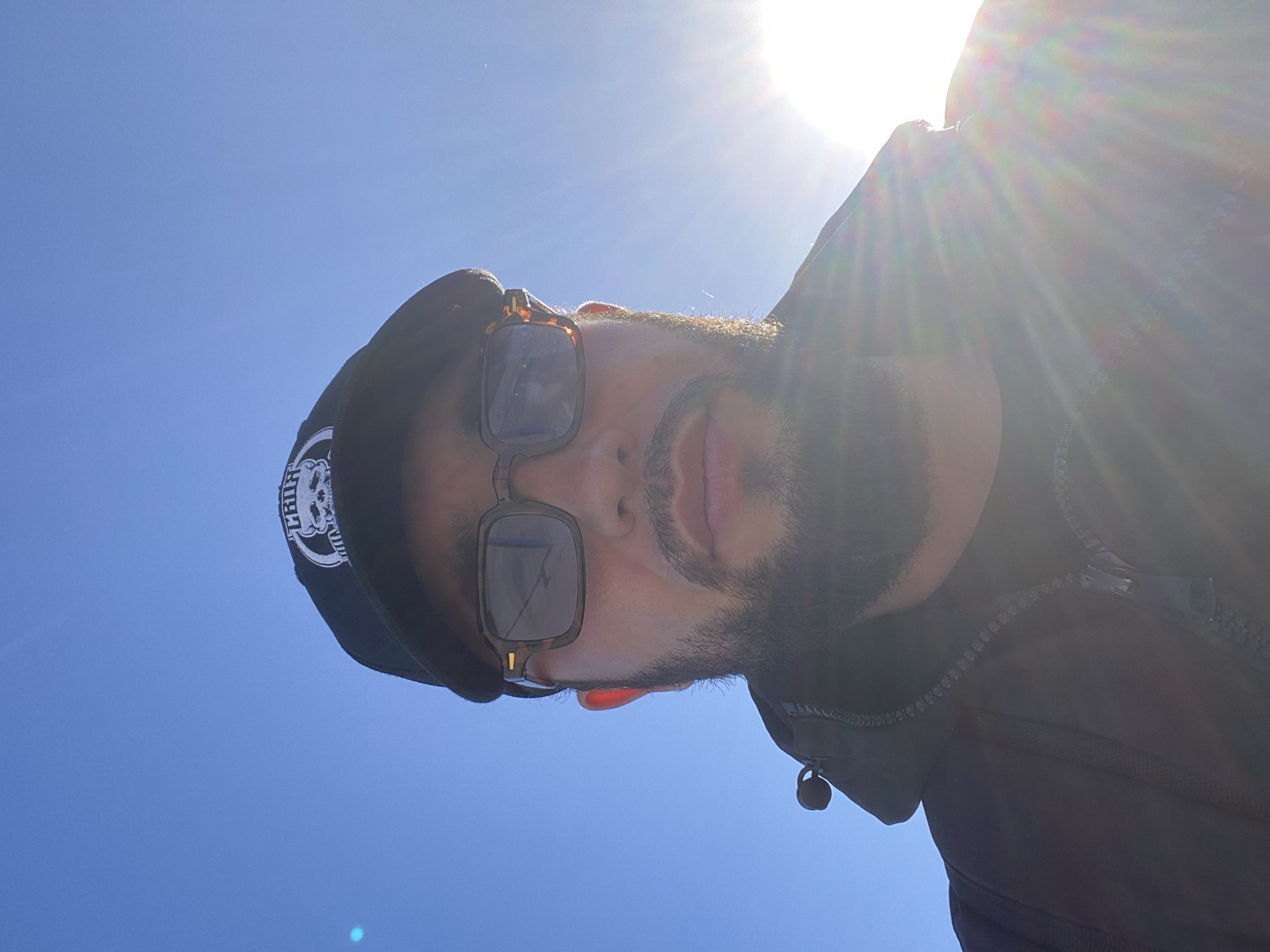 The image is a selfie taken outdoors against a bright blue sky, with the sun shining intensely and causing lens flare. The subject is a man wearing a black baseball cap with a white skull logo and patterned sunglasses. He has a beard and is wearing a dark jacket. The setting appears to be a sunny day, possibly with clear weather. There is no indication of the location being Koskolovo, Russia, or any specific urban environment.