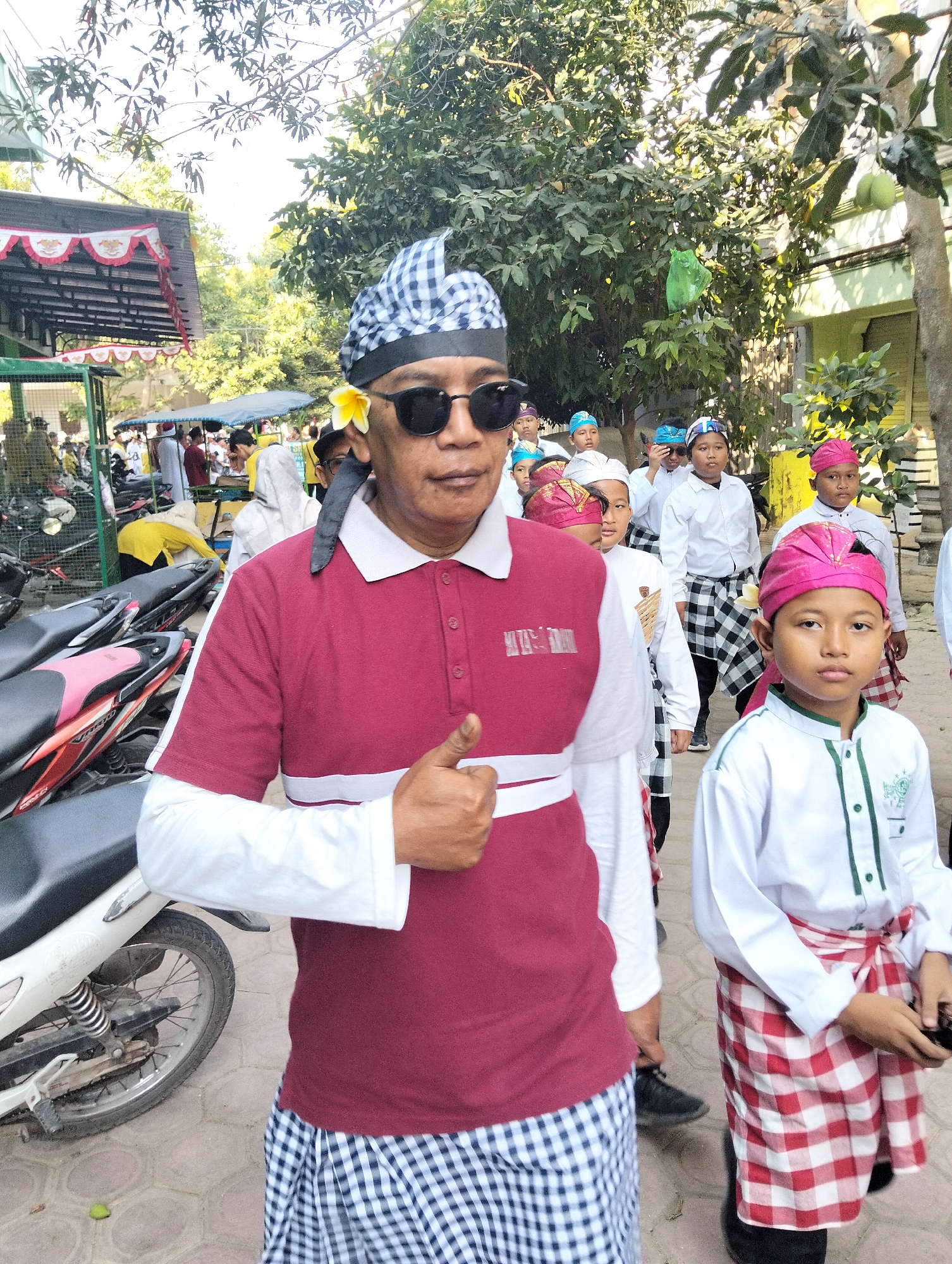 In Kranji, Indonesia, a man wearing a maroon and white polo shirt, sunglasses, and a blue and white checkered head covering, adorned with a yellow flower, gives a thumbs up while walking. He is accompanied by a group of boys dressed in white shirts and checkered sarongs, some wearing pink and blue head coverings. The scene appears to be outdoors, with trees and buildings in the background, and motorcycles parked nearby.