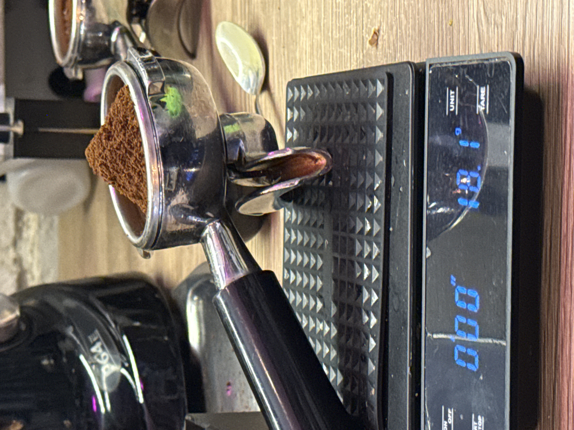 The image shows a close-up of a coffee preparation scene. In the foreground, a black digital scale displays "0.00" on the left and "18.1" with a "g" unit indicator on the right, suggesting it's measuring the weight of coffee grounds. A portafilter, filled with finely ground coffee, is placed on the scale's platform, which has a textured black surface. The portafilter is metallic with a black handle. Behind the scale and portafilter, a shiny black coffee grinder is partially visible, with "p64" printed on its side. A small, light-colored object, possibly a spoon or scoop, lies to the right of the portafilter on a wooden surface. The overall environment appears to be a kitchen or a cafe, with a focus on the process of weighing coffee for brewing. The image is well-lit, highlighting the details of the coffee and equipment. The provided image and its crops do not contain any information indicating the location of this scene within the city of Moscow, Russia.