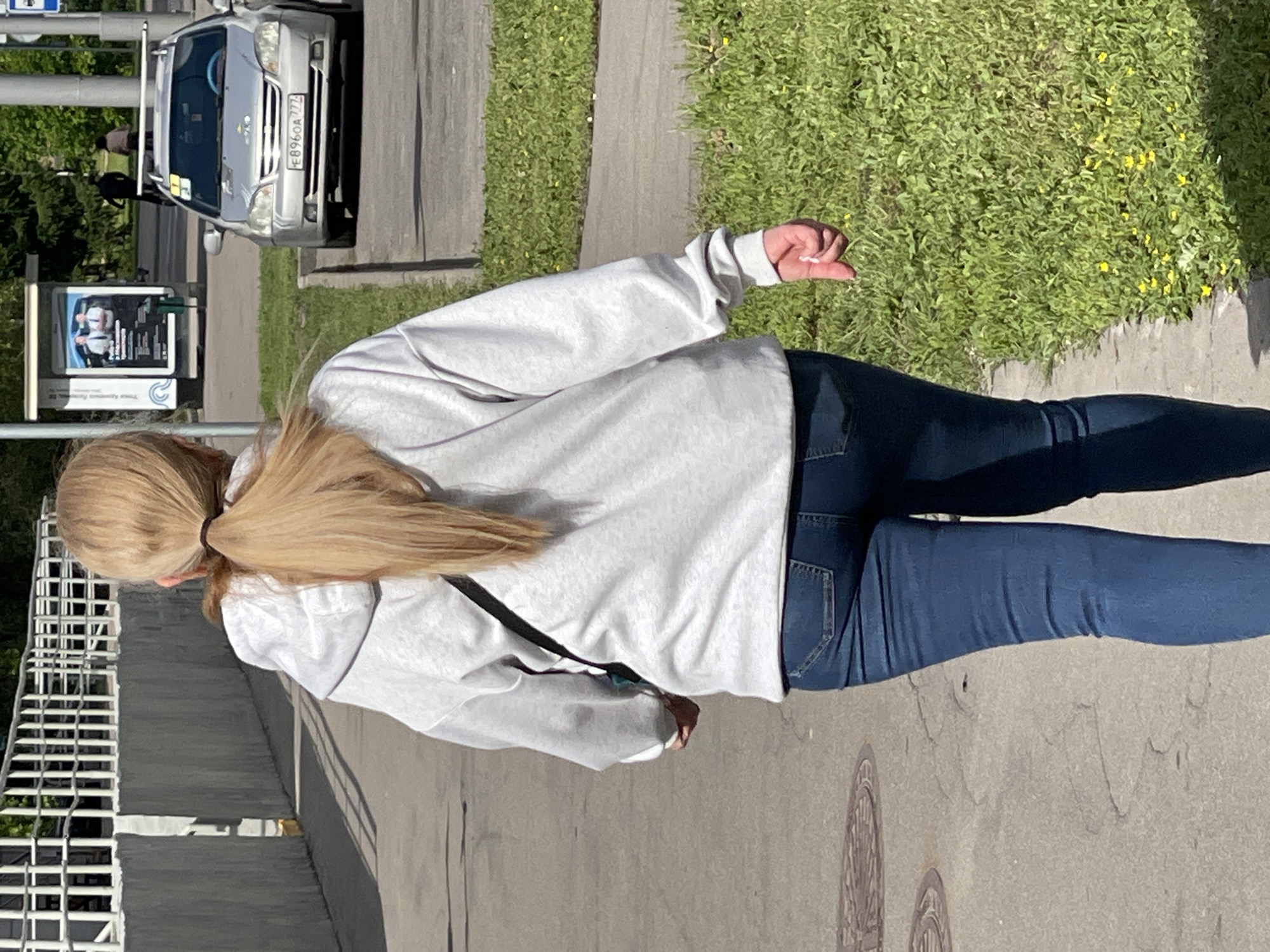A woman with blonde hair tied back in a ponytail walks on a sidewalk. She wears a light grey hoodie and blue jeans. She appears to be pointing with her right hand. A car with a license plate that reads "E896OA777" is parked on the side of the road. A sign, presumably advertising something, is visible near the car. The scene is located in Moscow, Russia.