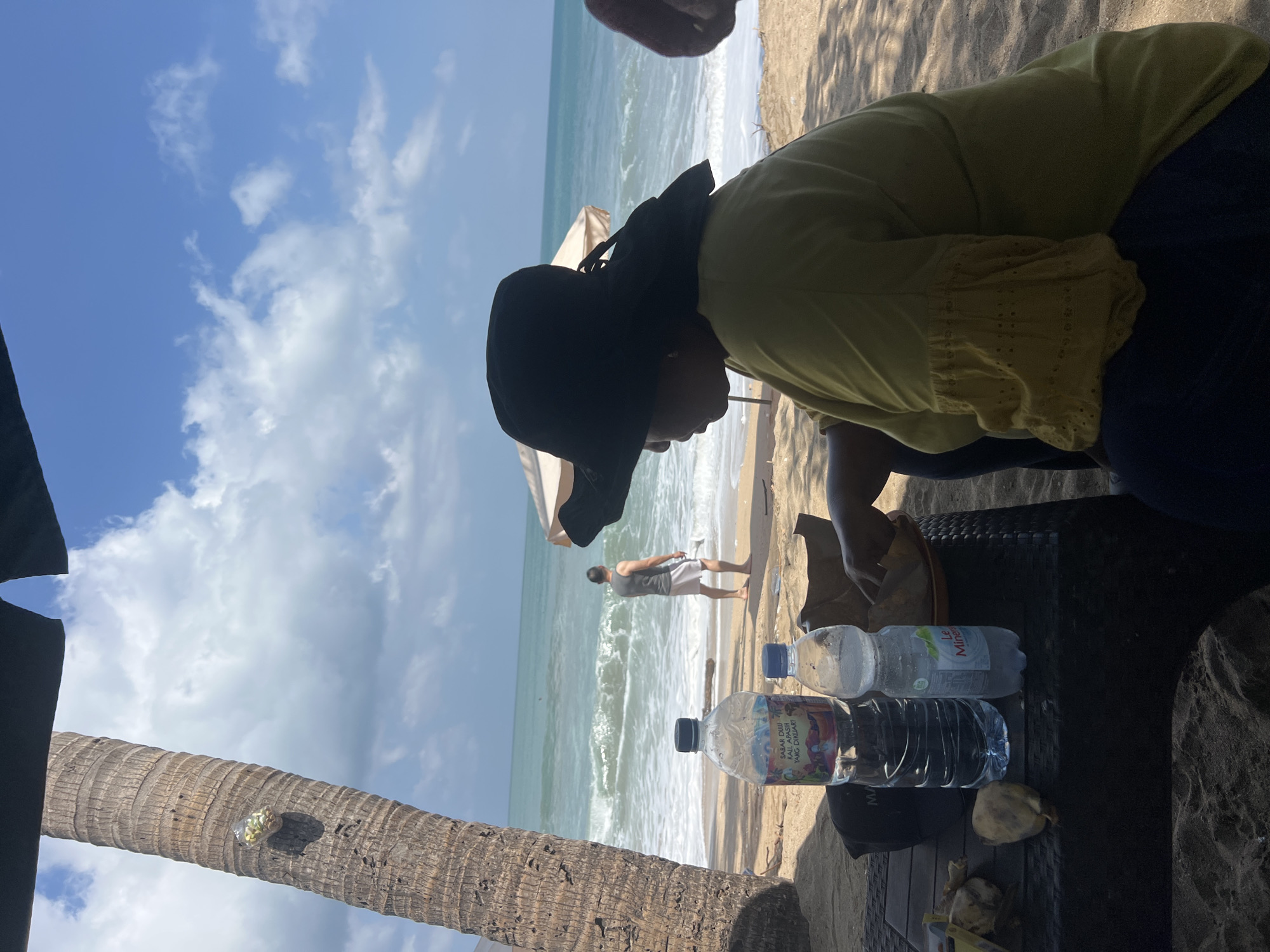The image depicts a beach scene in Kuta, Indonesia. In the foreground, a person wearing a black wide-brimmed hat and a yellow top is seated at a dark-colored table. On the table are two plastic bottles of water, one larger with a colorful label and a smaller one with a blue cap. There are also some food items, possibly fruit, and a brown paper bag. To the left of the table, a portion of a palm tree trunk is visible, with a small decorative item attached to it.
In the mid-ground, a man wearing a gray tank top and white shorts is standing on the beach, facing the ocean. The ocean has waves and a foamy shore. There are beach umbrellas in the background.
The overall environment is a sandy beach with a bright blue sky and scattered white clouds. The lighting suggests it is daytime, likely sunny.
