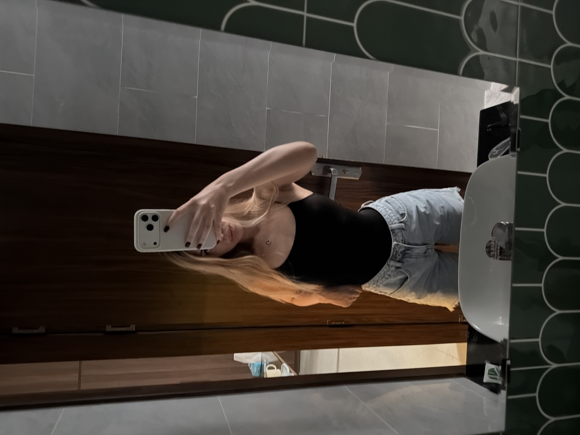 In a well-lit indoor environment, a person is taking a mirror selfie. The person, appearing to be female with blonde hair, is wearing a black tank top and light-wash denim shorts. They are holding a white smartphone to capture the reflection. The background includes a wooden door, tiled walls, and a portion of a sink basin. The overall aesthetic suggests a hotel room or a similar private dwelling, possibly in Đà Nẵng, Vietnam, judging by the typical hotel decor found in such tourist destinations.