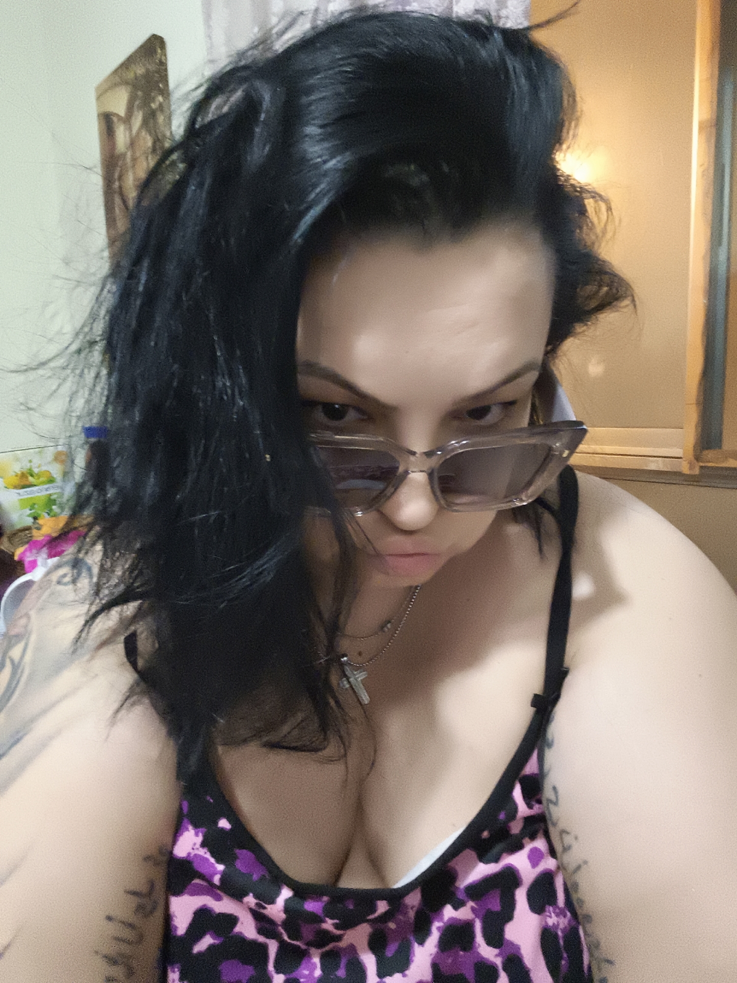 This is a medium shot, selfie-style photograph of a woman with dark, voluminous hair, wearing a leopard-print top and sunglasses. The woman is looking downwards at the camera, with her lips pursed. She is wearing a necklace with a cross pendant. Tattoos are visible on her left arm. The background is softly lit and appears to be an indoor setting, possibly a room with a window and a framed picture on the wall. There is no visible indication of the city of Komotini, Greece in the image.