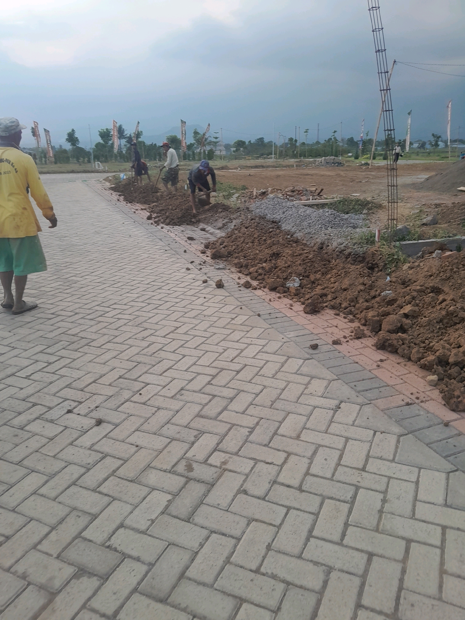 The image depicts a construction site in Pakisaji, Indonesia. The foreground is dominated by a paved walkway made of light-colored interlocking bricks laid in a herringbone pattern. A person wearing a yellow shirt and green shorts is walking away from the camera on this walkway. In the mid-ground, several workers are engaged in digging and moving dirt with shovels. Piles of soil and gravel are visible along the edge of the walkway. In the background, construction materials, including rebar for a pillar, are present. Several flags are planted in the distance, and trees and hills can be seen under an overcast sky, suggesting a rural or semi-urban environment. The overall scene is one of ongoing construction and land preparation.