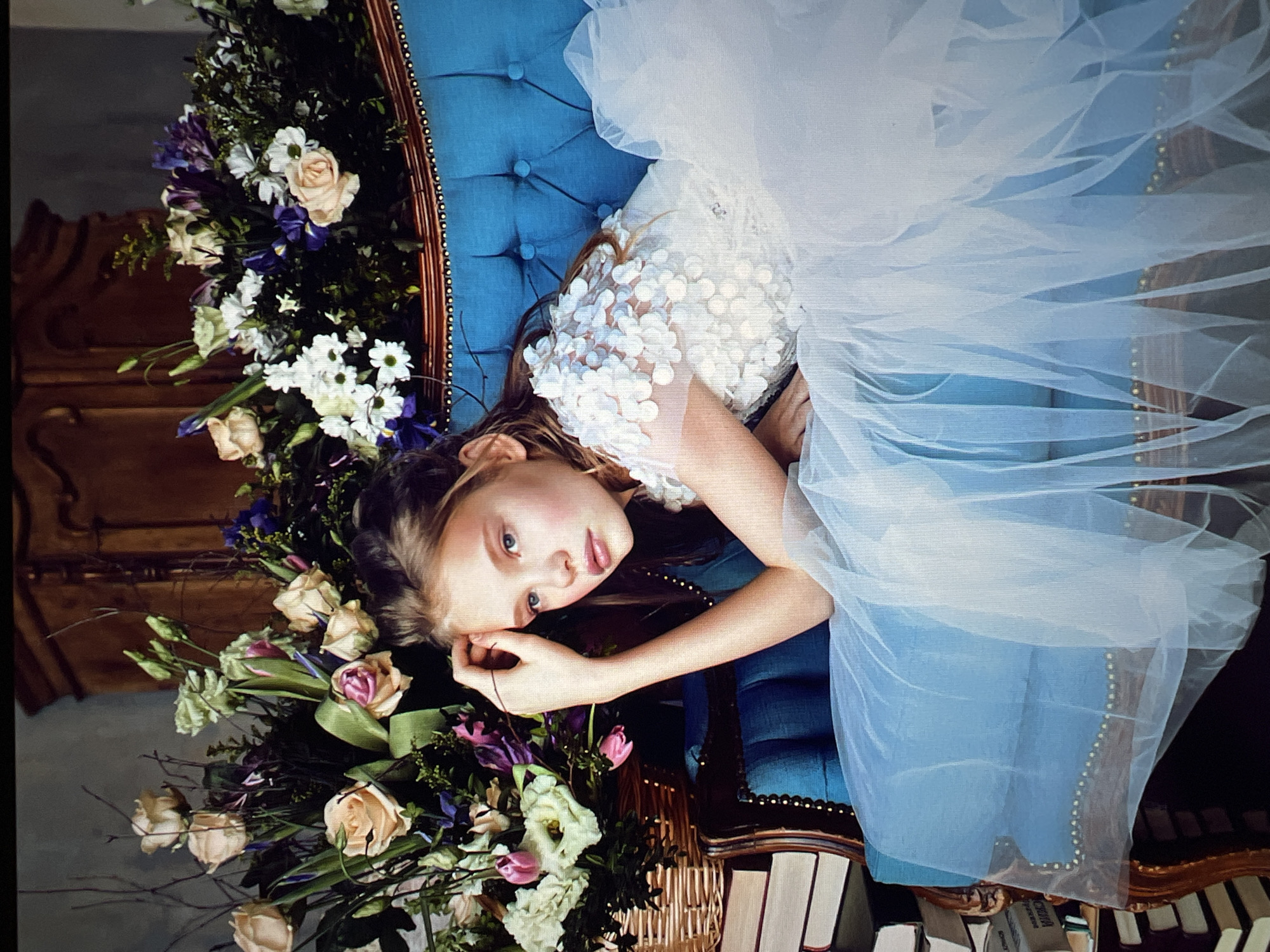 An elegant, blonde-haired young girl with striking blue eyes is the central subject of this studio portrait. She is posed reclining on a plush, tufted, bright blue velvet sofa, her head resting on her left hand. Her gaze is directed forward with a thoughtful expression. She is adorned in a delicate white dress, featuring a sheer, ethereal overlay on the skirt and intricately embellished with floral appliques on the bodice and sleeves.
Behind and around her, a lavish arrangement of fresh flowers creates a vibrant backdrop. The bouquet is a rich mix of colors and textures, including pale peach roses, delicate white daisies, purple irises, and pink tulips, interspersed with lush greenery and dark, dramatic branches. The flowers are partially contained within a woven wicker basket to the left of the frame.
To the left of the sofa, a stack of old books is visible, suggesting a scholarly or fairytale-inspired setting. The background is a muted, textured grey wall, with a glimpse of ornate wooden furniture, possibly an armoire, further enhancing the sophisticated aesthetic of the scene. The lighting appears to be soft and diffused, highlighting the subject and her surroundings with a gentle glow. The overall impression is one of delicate beauty and timeless grace.