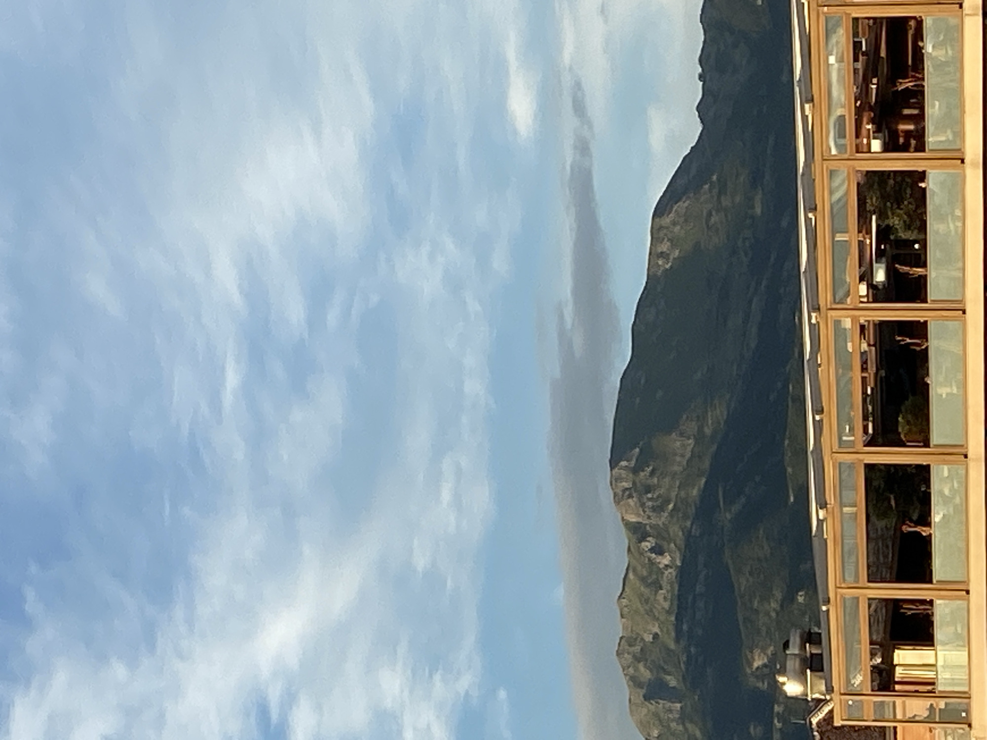 A wide shot captures the mountainous landscape of Kovačko Polje, Montenegro. The sky is a pale blue, dotted with wispy white clouds. In the mid-ground, a lush green mountain range dominates the scene, its rugged peaks catching the sunlight. In the foreground, a modern building with a glass facade and wooden accents is visible. The building's interior, partially visible through the glass, shows sparse furnishings and some greenery.