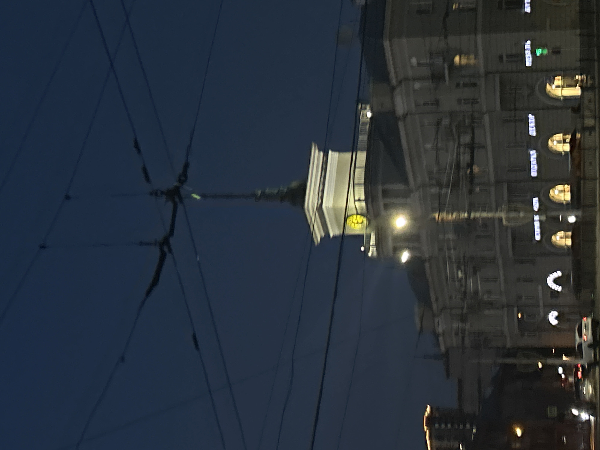 The image depicts a street scene at dusk in Barnaul, Russia. Dominating the upper portion of the frame are numerous overhead wires, likely for public transportation, crisscrossing the dark blue sky. Below the wires, a distinctive building with a tower featuring a clock is visible. This structure appears to be a prominent landmark. The building's facade is adorned with multiple windows, some illuminated, suggesting indoor activity.
In the lower part of the image, several vehicles are present on the street, with headlights and taillights indicating movement or stationary positions. The street itself appears to be paved. The overall environment suggests an urban setting during the evening hours, with artificial lights illuminating parts of the street and buildings. The buildings in the background, though less distinct, reinforce the urban context. The presence of streetlights and illuminated windows contributes to a sense of a city that is active even as daylight fades. The image captures a moment in time on a street in Barnaul, Russia, characterized by its infrastructure and architecture under the twilight sky.