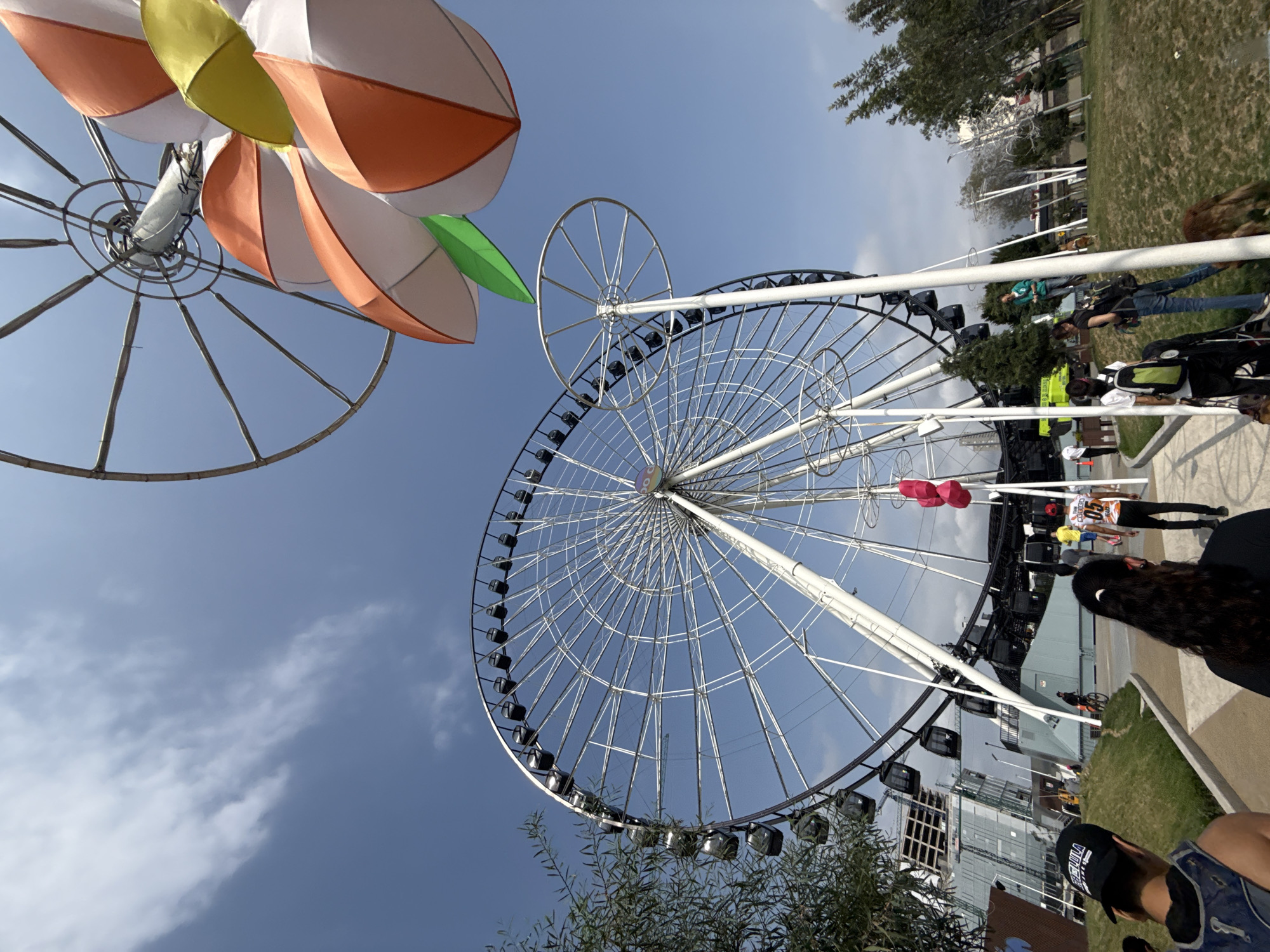 The image captures a Ferris wheel in Puebla, Mexico. A floral-themed structure is on the left side. A large Ferris wheel with passenger cars dominates the scene. The lower portion of the frame shows trees and parts of buildings. People are present, with some visible along a walkway. The sky is blue with clouds.